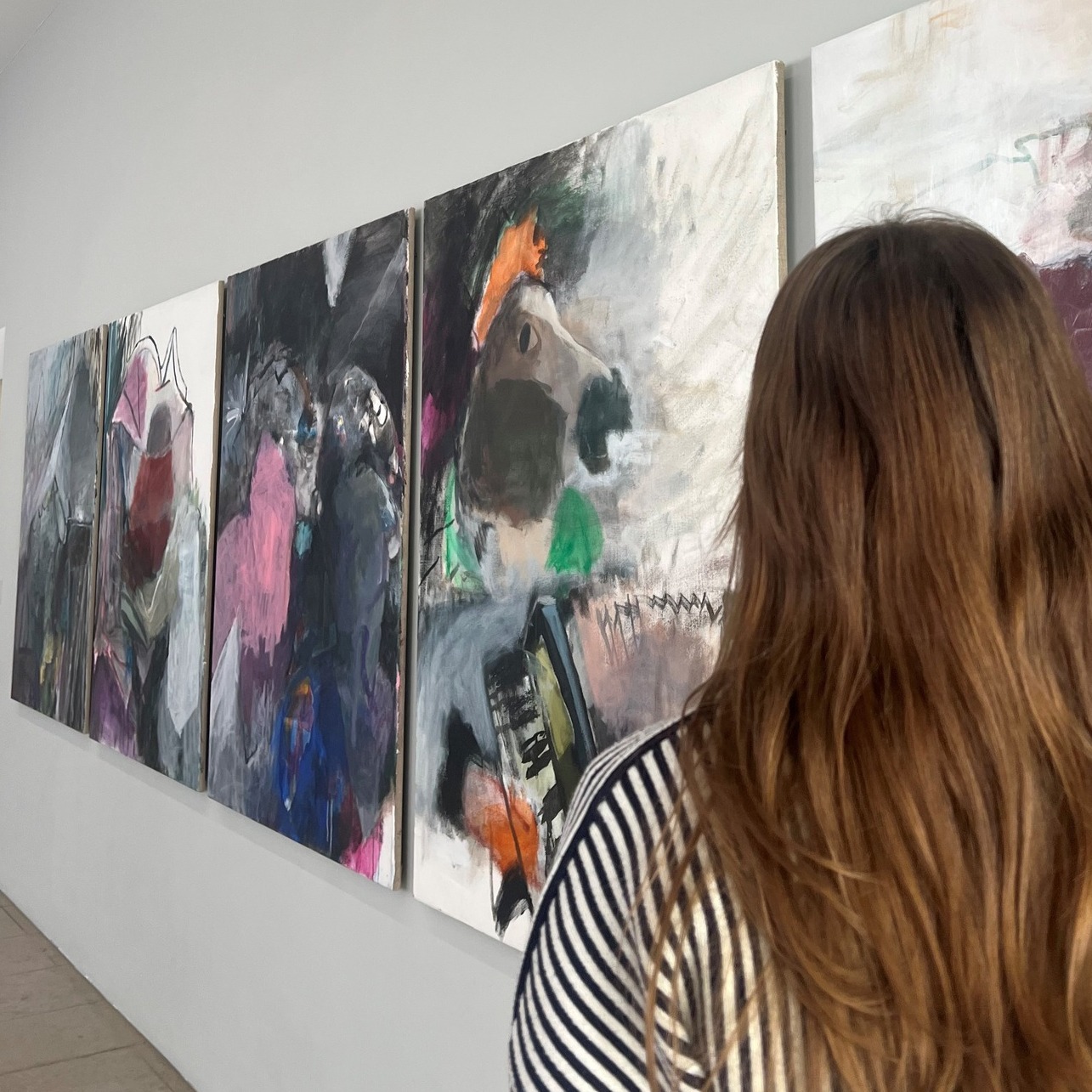 Dieser Alternativtext wurde KI-generiert: Eine Person betrachtet eine Reihe abstrakter Gemälde an einer Wand. Die Kunstwerke sind in verschiedenen Farben und Stilrichtungen gehalten.