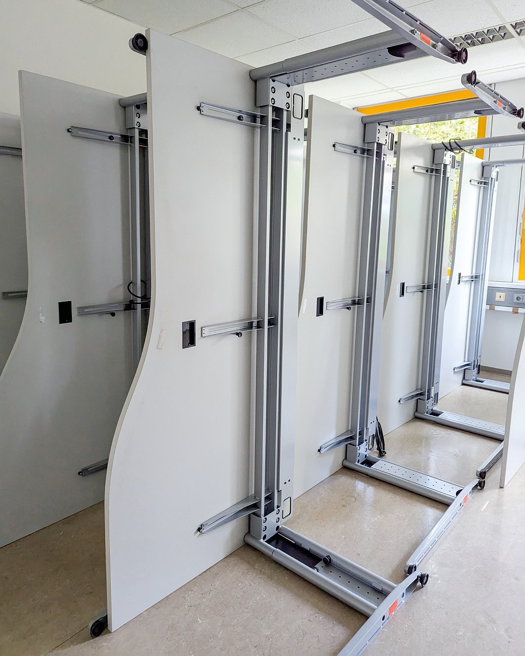 Dieser Alternativtext wurde KI-generiert: Mehrere weiße Paneele sind an Metallschienen montiert und stehen auf Rädern. Die Konstruktion wirkt wie eine Art Raumteiler oder mobile Wand.