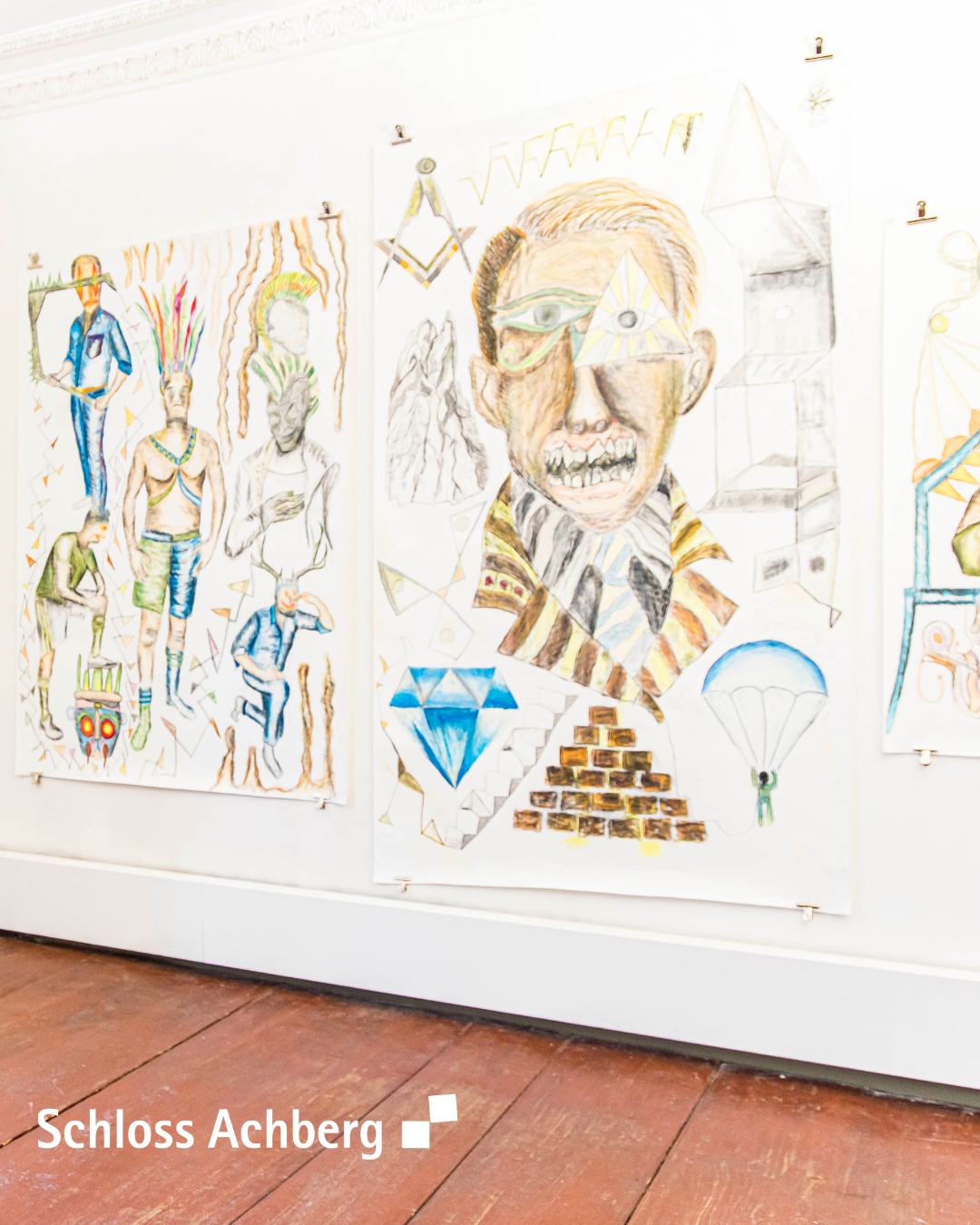 Dieser Alternativtext wurde KI-generiert: Eine Wand zeigt mehrere farbenfrohe, abstrakte Zeichnungen und Gemälde. Im Vordergrund befindet sich ein Porträt mit einem auffälligen Diamanten darunter.