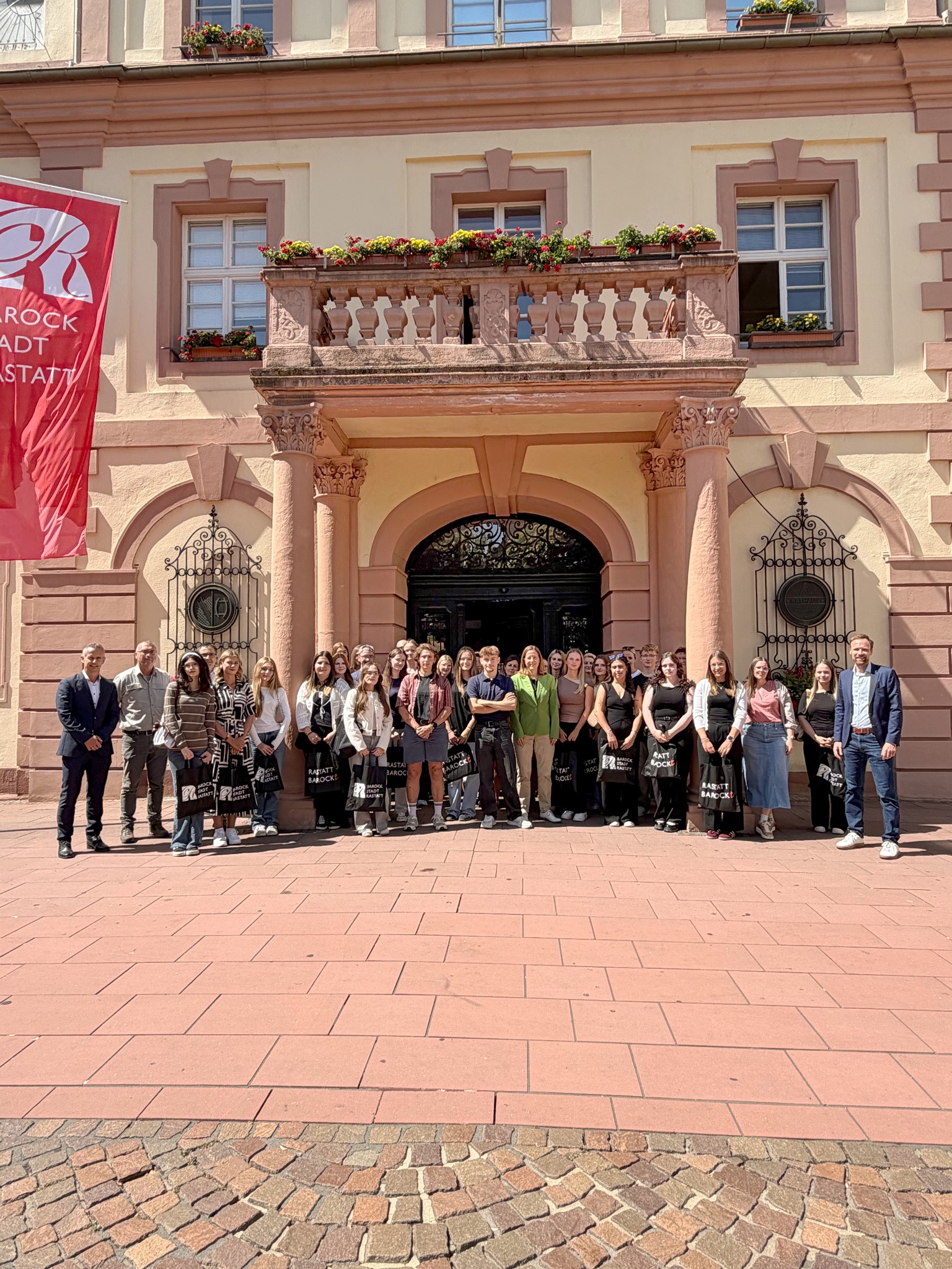 Eine große Gruppe von Menschen steht vor dem Eingang eines historischen Gebäudes mit rosa Fassade und Blumenbalkon. Viele Personen halten schwarze Taschen mit der Aufschrift "RASTATT BAROCK". Links im Bild hängt eine rote Flagge mit dem Schriftzug "BAROCK STADT RASTATT". (KI-Alternativtext)