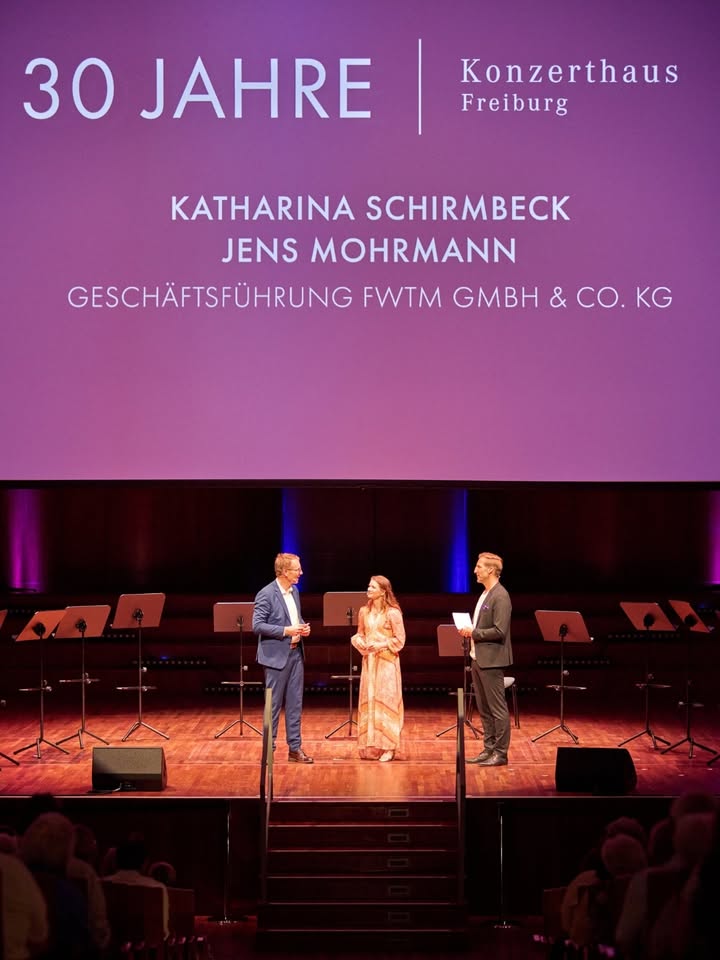 Drei Personen stehen auf einer Bühne vor einem großen violetten Bildschirm, auf dem „30 JAHRE Konzerthaus Freiburg“ sowie die Namen „KATHARINA SCHIRMBECK“ und „JENS MOHRMANN“ mit der Funktion „GESCHÄFTSFÜHRUNG FWTM GMBH & CO. KG“ zu lesen sind. Im Hintergrund der Bühne stehen mehrere Notenständer, während im unteren Bildrand die Köpfe des Publikums sichtbar sind. (KI-Alternativtext)