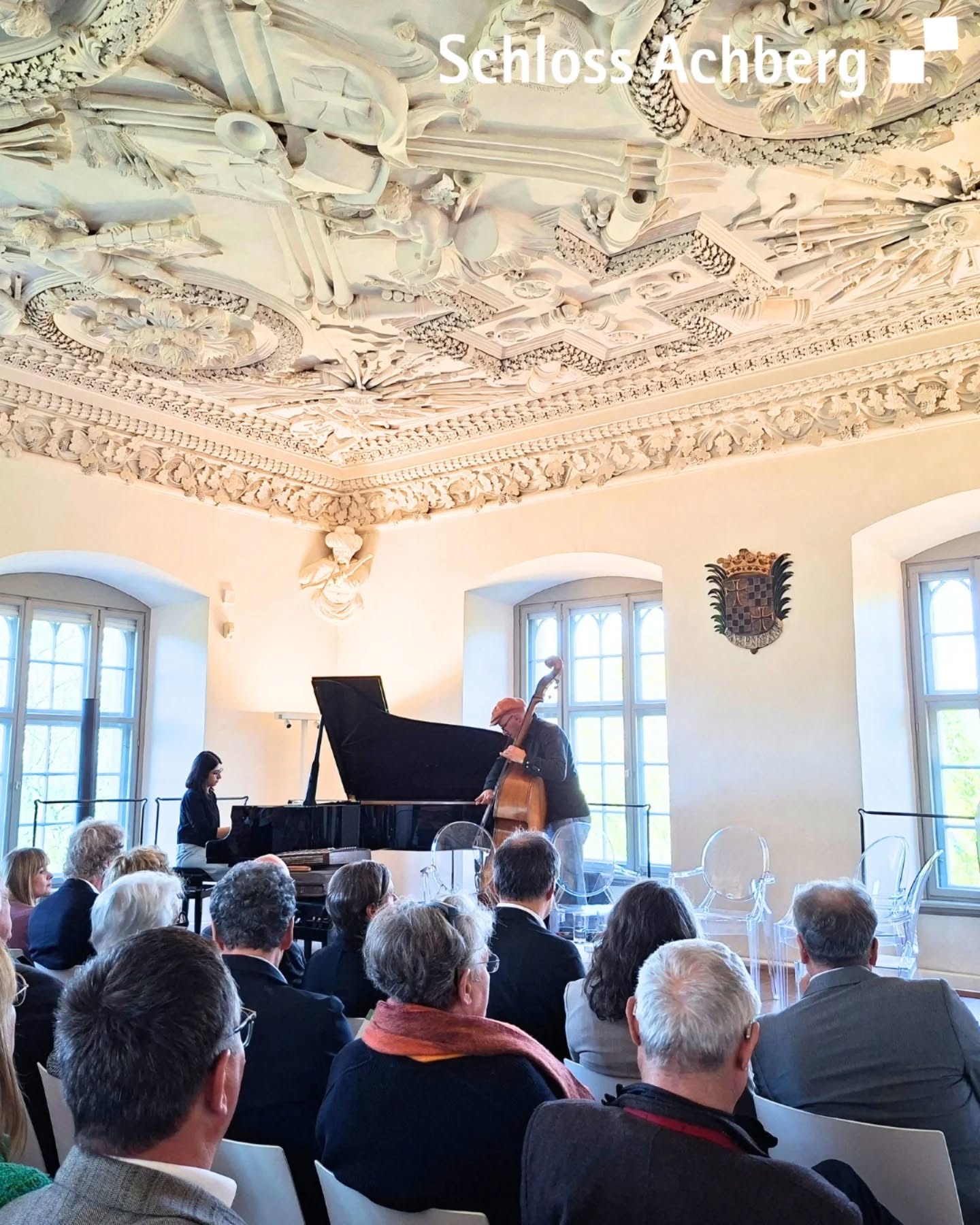 Dieser Alternativtext wurde KI-generiert: Innenansicht eines Raumes mit aufwendiger Stuckdecke. Eine musikalische Darbietung mit Piano und Kontrabass findet vor einem Publikum statt.