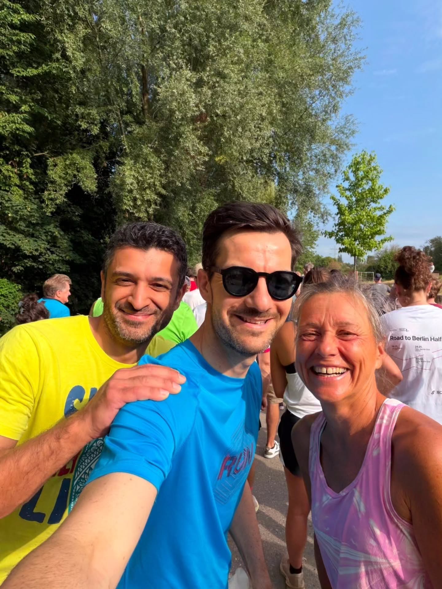Drei Personen posieren lächelnd für ein Selfie im Freien bei Sonnenschein. Die Person in der Mitte trägt ein blaues T-Shirt mit der Aufschrift „RUN“ und eine Sonnenbrille, flankiert von einer Person in einem gelben Shirt links und einer in einem rosa Top rechts. Im Hintergrund sind Bäume und weitere Personen zu sehen, darunter eine mit einem Shirt, auf dem „Road to Berlin Half“ steht. (KI-Alternativtext)