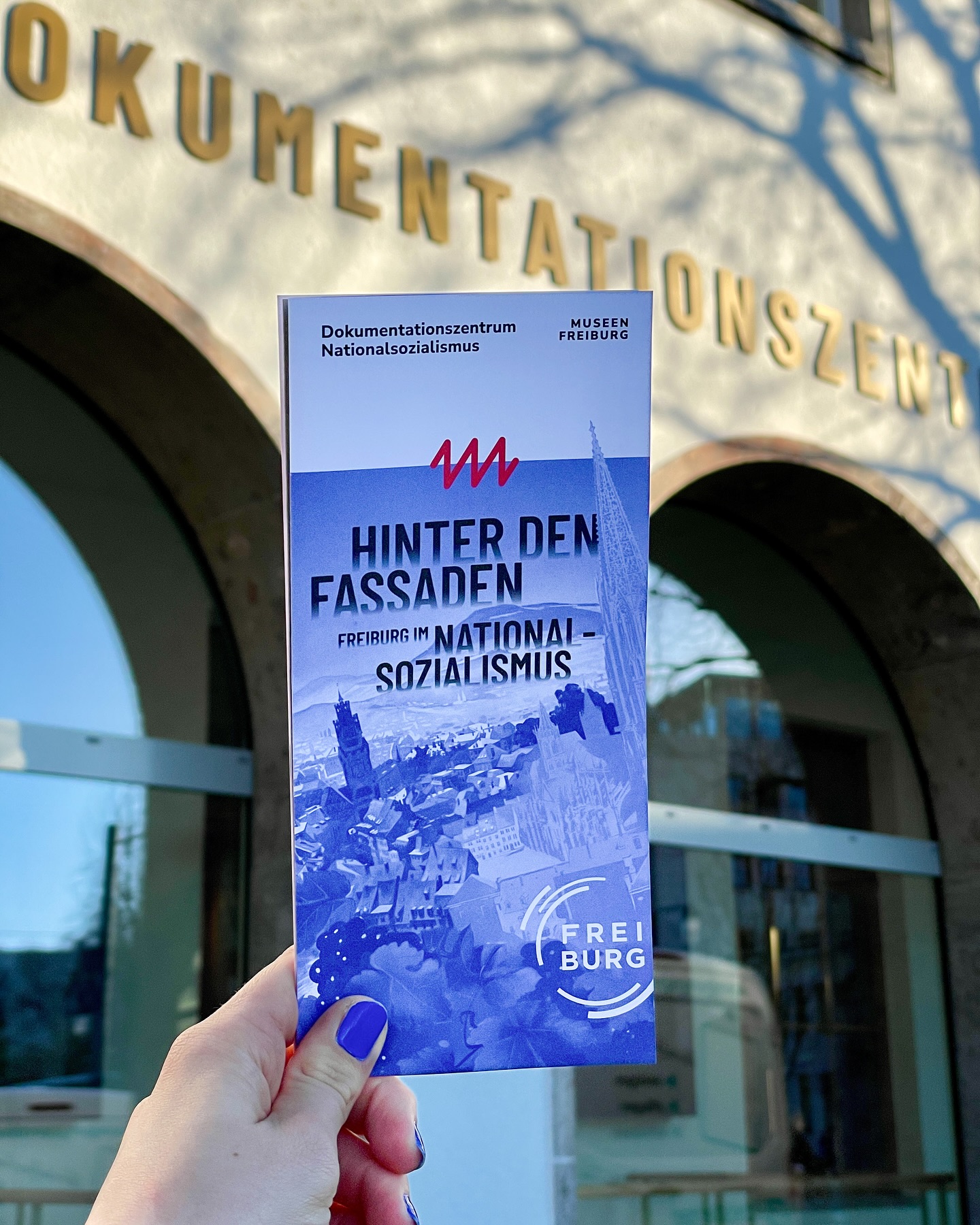 Hand hält einen blauen Flyer vor einem Gebäude. Der Flyer zeigt ein Stadtbild und den Titel 