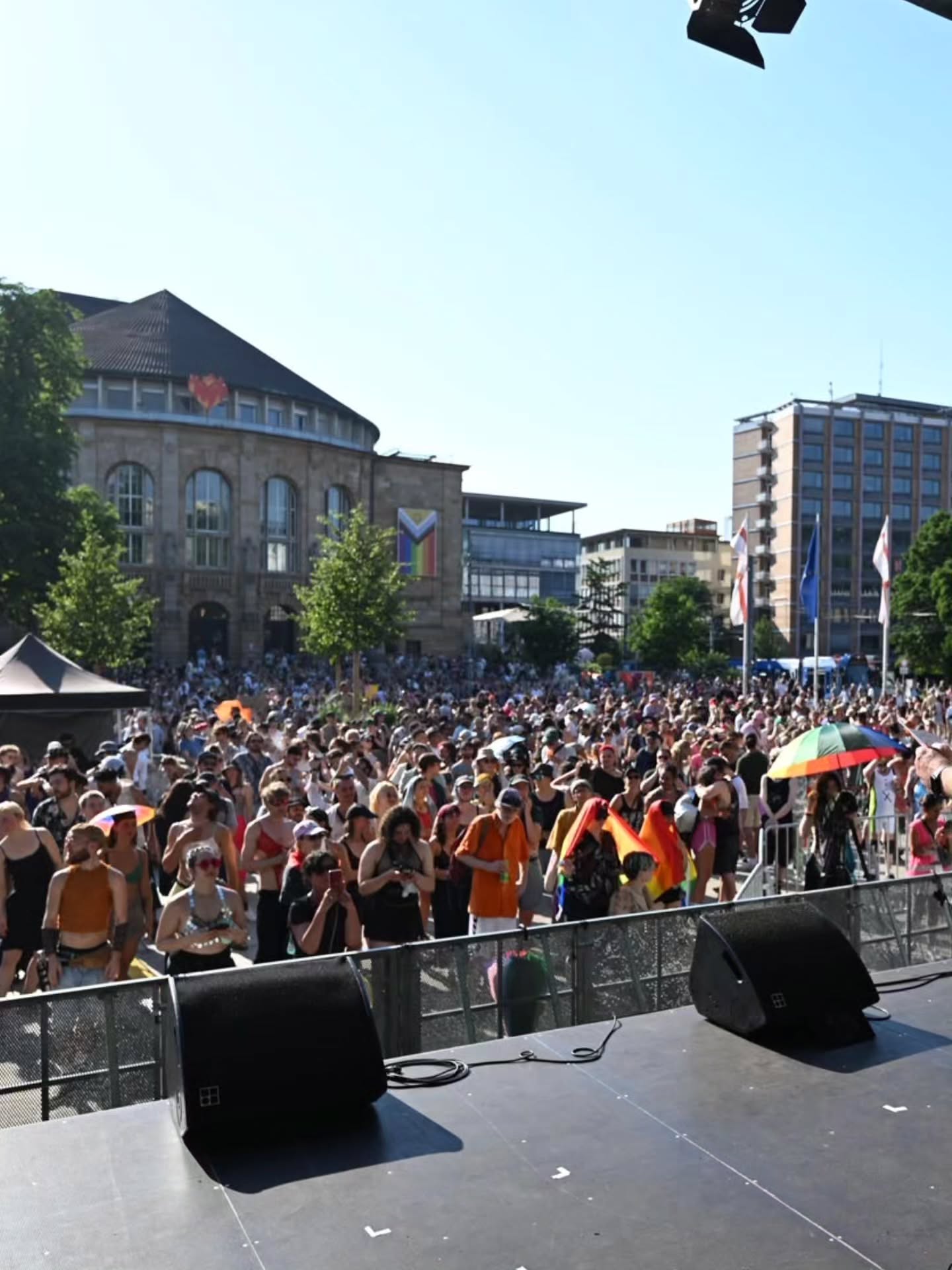 Blick von einer Bühne auf eine große Menschenmenge bei einem sonnigen Straßenfest. Im Hintergrund ein großes Gebäude mit Regenbogenflagge und Herzsymbol, einige Personen halten Regenbogenfahnen oder -schirme. (KI-Alternativtext)