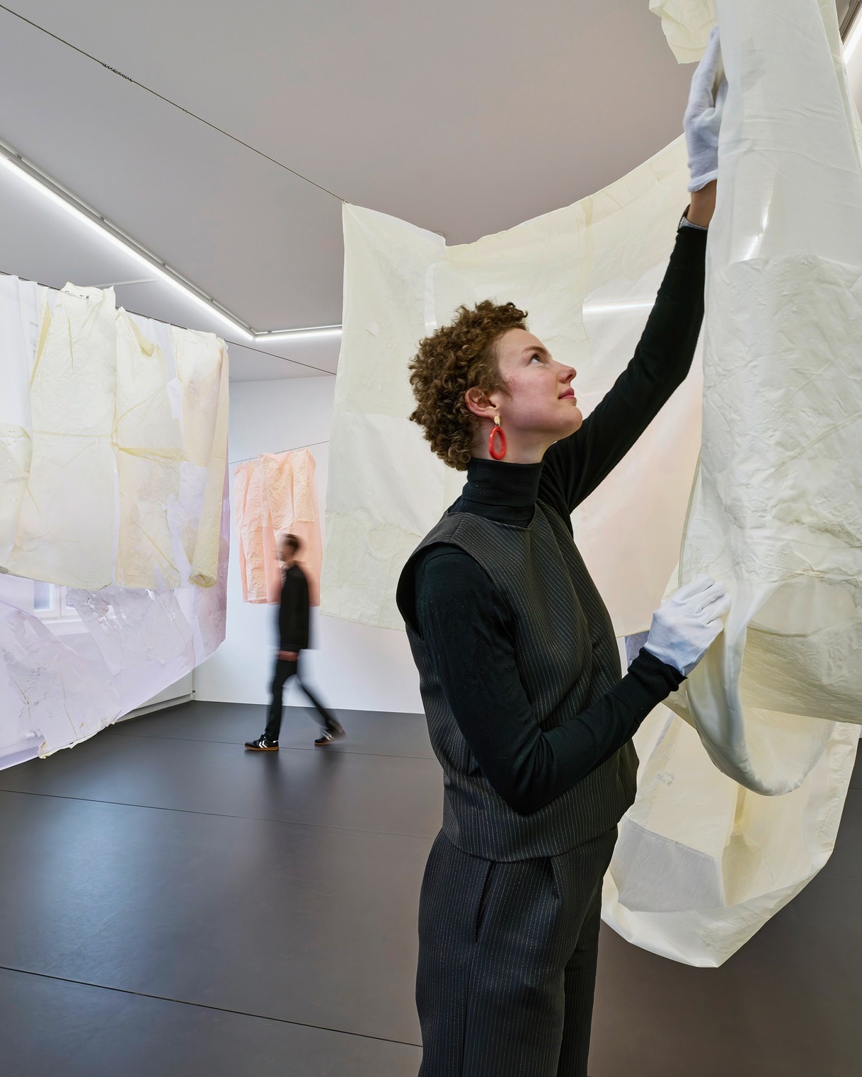 Dieser Alternativtext wurde KI-generiert: Eine Person mit lockigem Haar trägt weiße Handschuhe und bearbeitet ein großes, beiges Stoffstück in einem hellen Raum. Im Hintergrund ist eine weitere Person zu sehen, die sich in dem Raum bewegt.