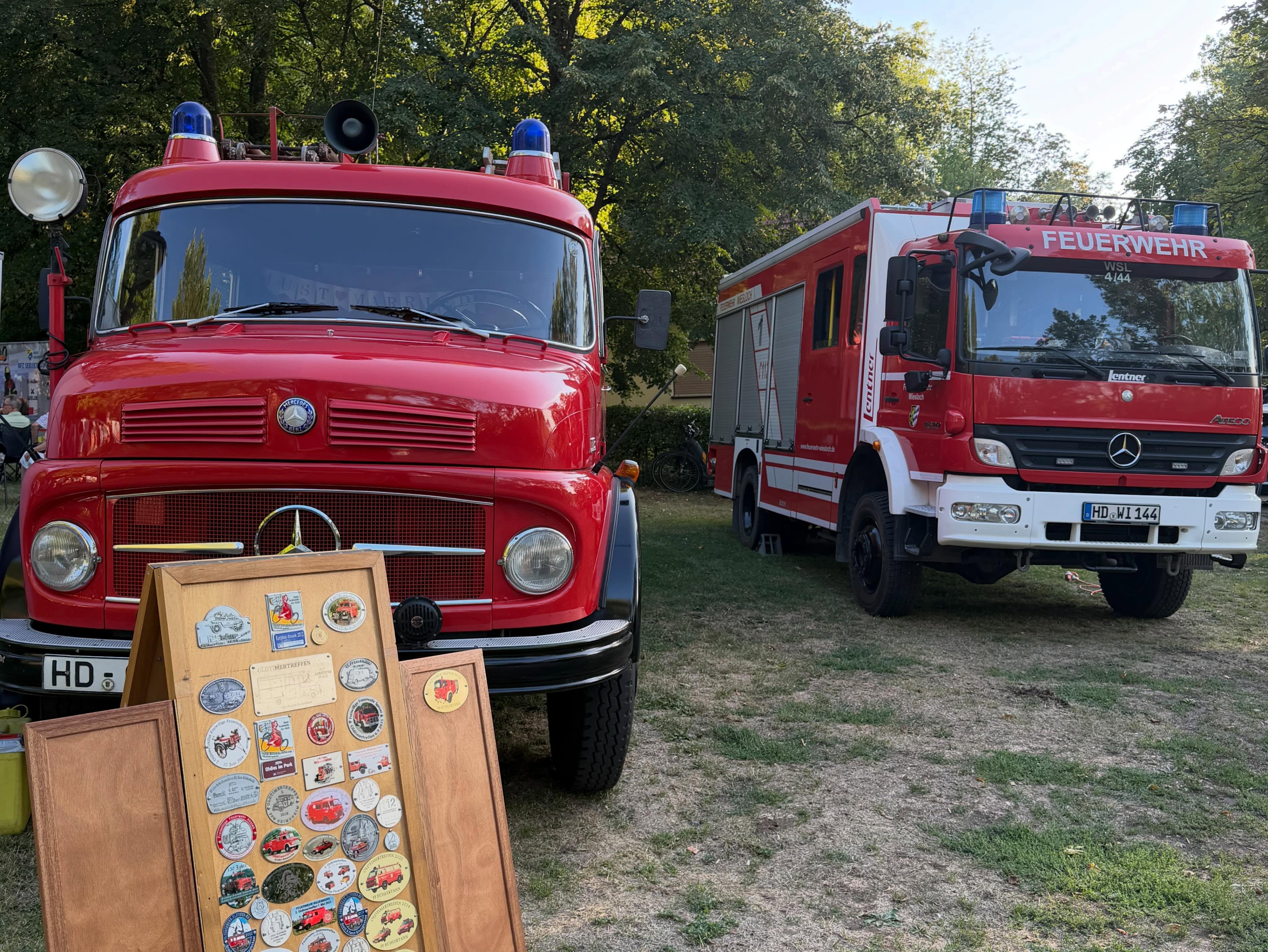 Zwei rote Feuerwehrfahrzeuge stehen auf einer Wiese vor einem Hintergrund aus Bäumen. Im Vordergrund steht ein klassisches rotes Fahrzeug mit einem Holzschild voller Aufkleber davor, während rechts daneben ein modernes rotes und weißes Fahrzeug mit der Aufschrift Feuerwehr parkt. (KI-Alternativtext)