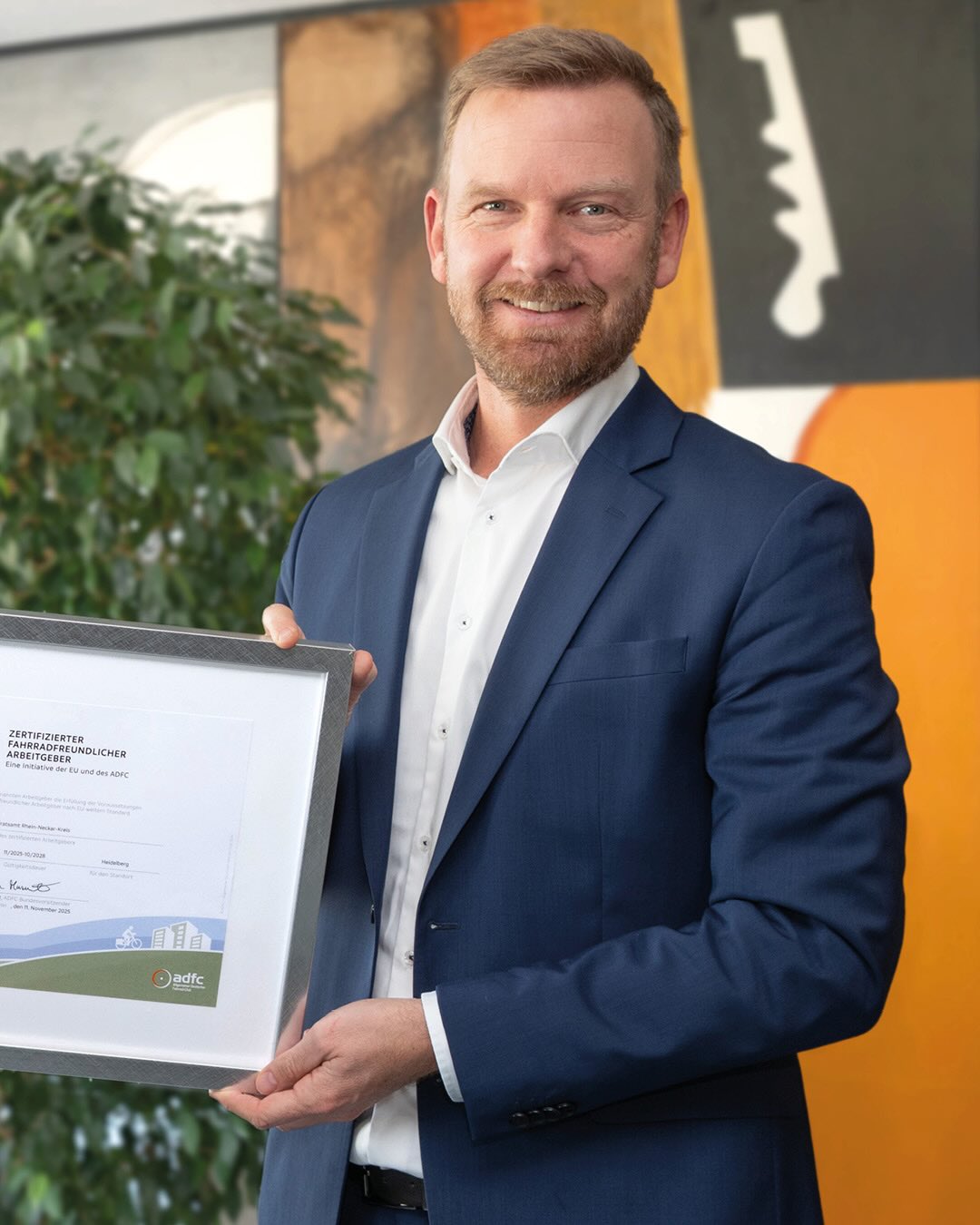 Dieser Alternativtext wurde KI-generiert: Eine Person in einem Anzug hält ein Zertifikat in der Hand. Im Hintergrund sind Bäume und ein Gebäude zu sehen.