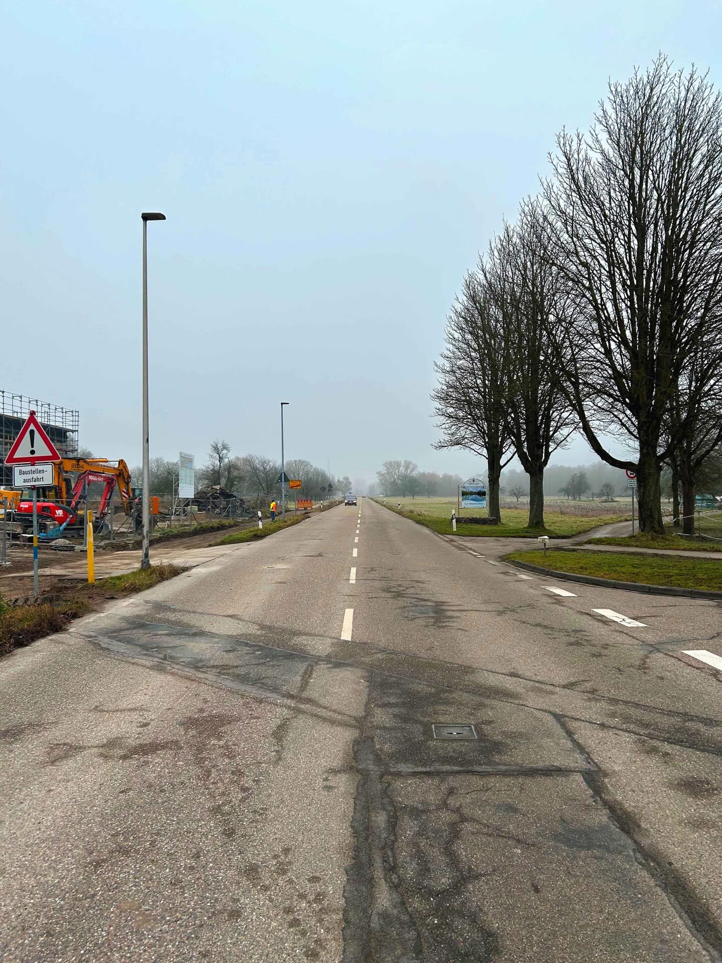 Dieser Alternativtext wurde KI-generiert: Eine lange, gerade Straße führt in die Ferne, gesäumt von Bäumen und Feldern. Ein Warnschild und Baumaschinen sind am Straßenrand zu sehen.