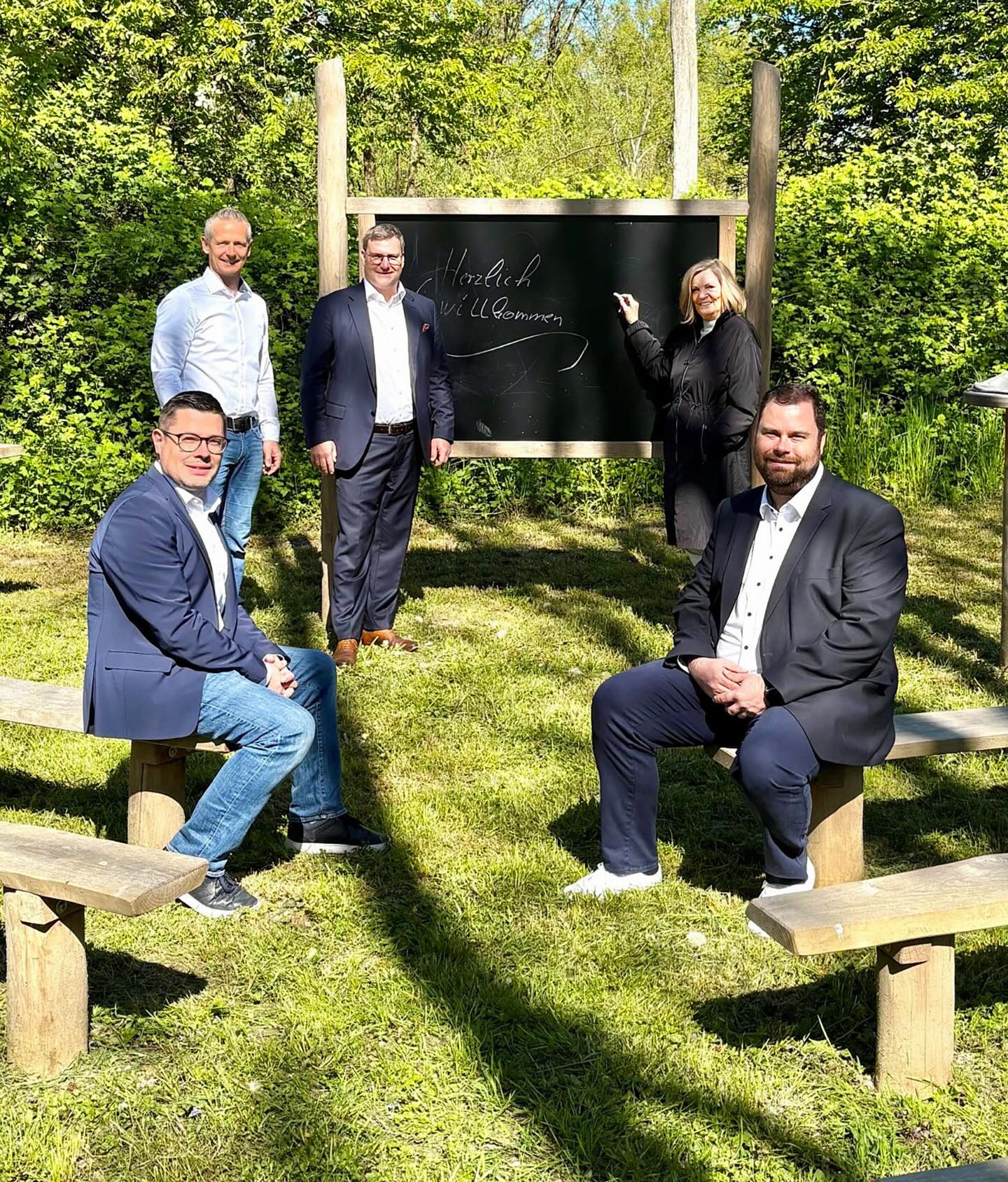 Dieser Alternativtext wurde KI-generiert: Eine Gruppe von Menschen steht und sitzt vor einer Tafel in einer Gartenlandschaft. Die Aufschrift auf der Tafel ist in Weiß gehalten.