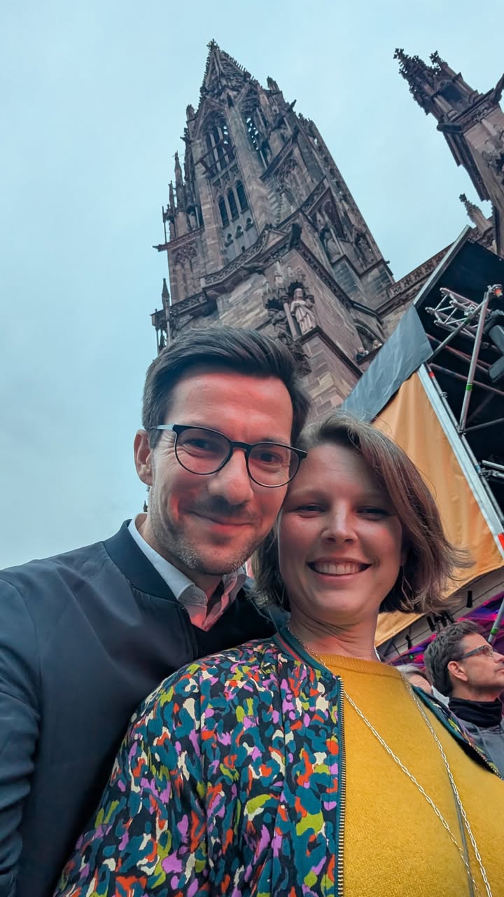 Zwei Personen lächeln in die Kamera, im Hintergrund ein hoher, gotischer Kirchturm und ein Teil einer Bühne mit Gerüst. Eine Person trägt eine Brille und eine dunkle Jacke, die andere eine bunt gemusterte Jacke über einem gelben Oberteil. (KI-Alternativtext)