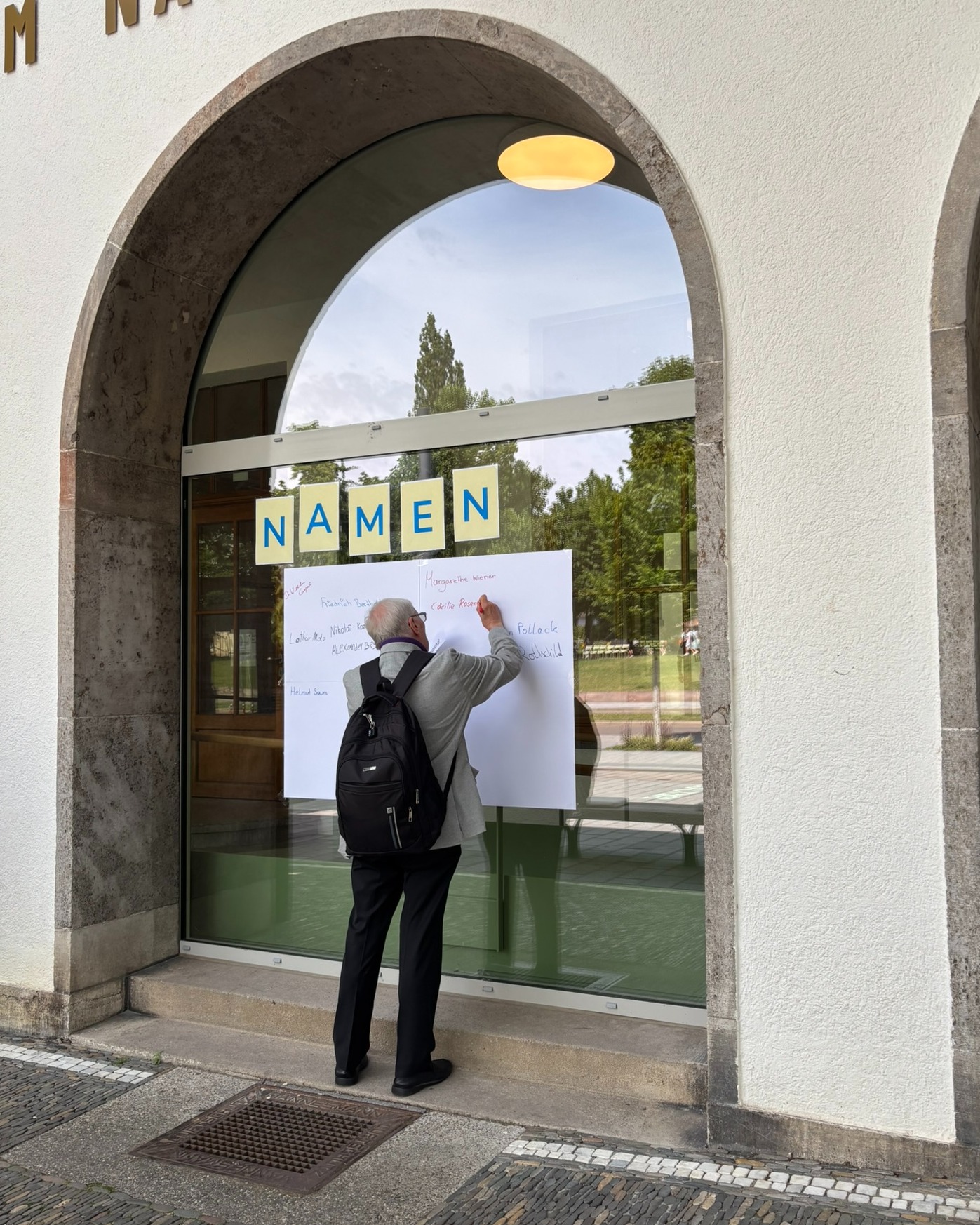 Eine Person mit Rucksack steht vor einem großen Fensterbogen und schreibt auf ein weißes Blatt Papier, das am Glas befestigt ist. Über dem Papier kleben gelbe Zettel, die das Wort "NAMEN" bilden, während auf dem Blatt bereits weitere handschriftliche Namen zu sehen sind. (KI-Alternativtext)