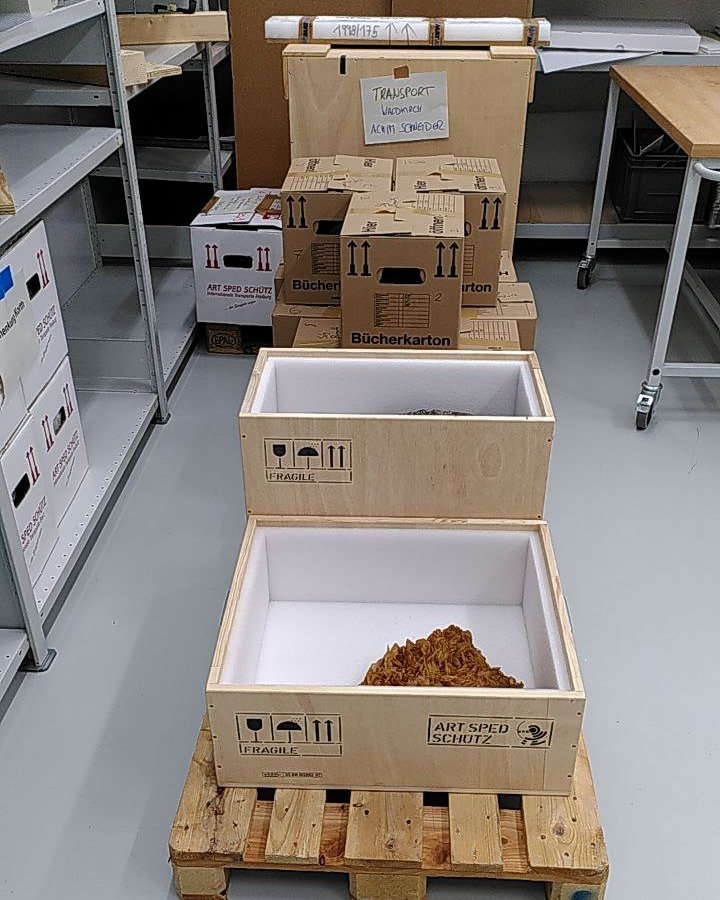 Dieser Alternativtext wurde KI-generiert: Mehrere Holzkisten und Kartons sind in einem Lagerraum zu sehen. Eine offene Kiste enthält ein bräunliches, unregelmäßiges Material.