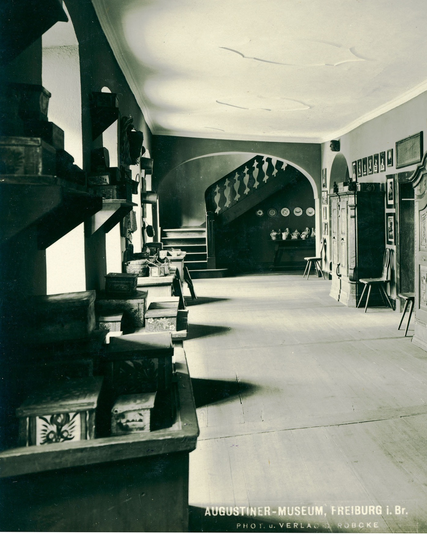 Dieser Alternativtext wurde KI-generiert: Schwarz-weißes Foto einer langen, hellen Halle mit einer Treppe im Hintergrund. Entlang der Wände stehen Möbelstücke und dekorative Gegenstände.