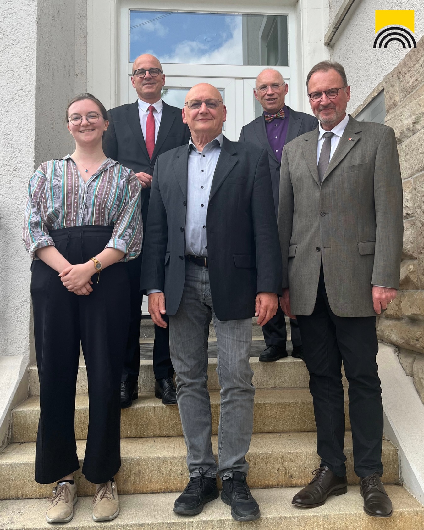 Aufnahme von fünf Personen vor einer Tür, die auf einer Steintreppe stehen. Die Personen tragen formelle Kleidung.