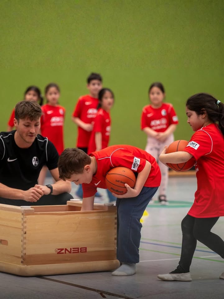 Dieser Alternativtext wurde KI-generiert: Mehrere Personen stehen und sitzen in einer Sporthalle. Ein Basketball wird zwischen zwei Personen weitergegeben, während andere zuschauen.