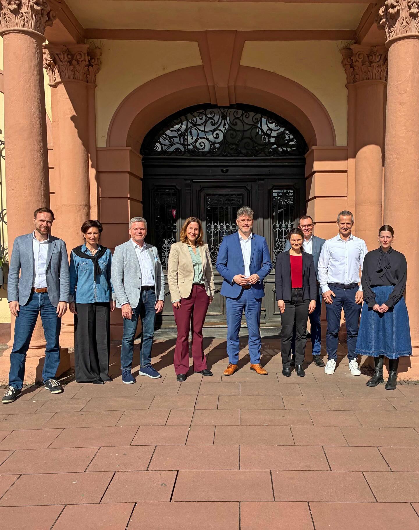 Dieser Alternativtext wurde KI-generiert: Eine Gruppe von Menschen steht vor einem Gebäude mit einem verzierten Eingangsbereich. Das Bild ist farbig und zeigt eine Außenaufnahme bei Tageslicht.