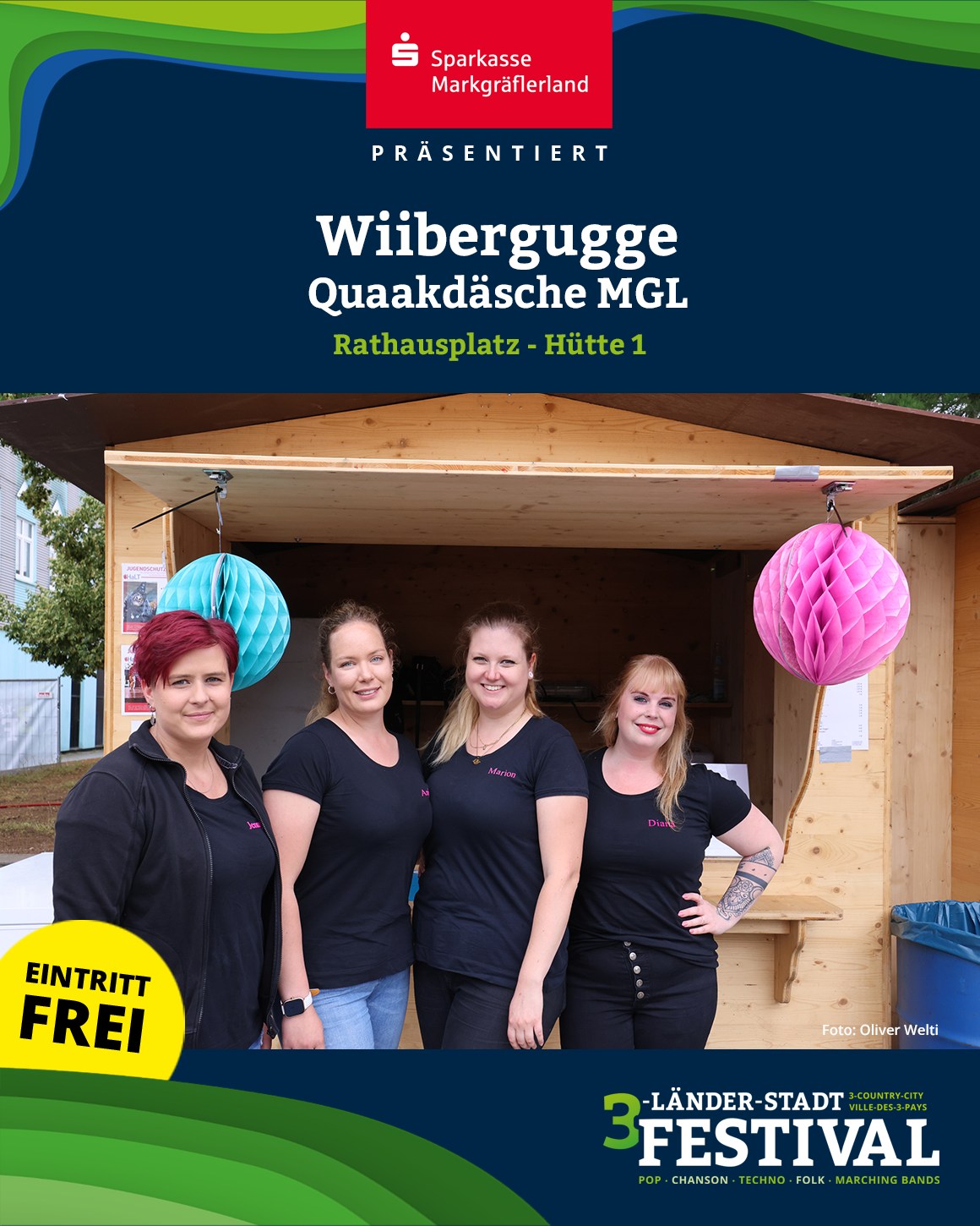 Eine Gruppe von vier Personen steht vor einem Holzgebäude, das mit blauem und pinkem Papier geschmückt ist. Im Vordergrund steht in gelber Schrift "Eintritt Frei".