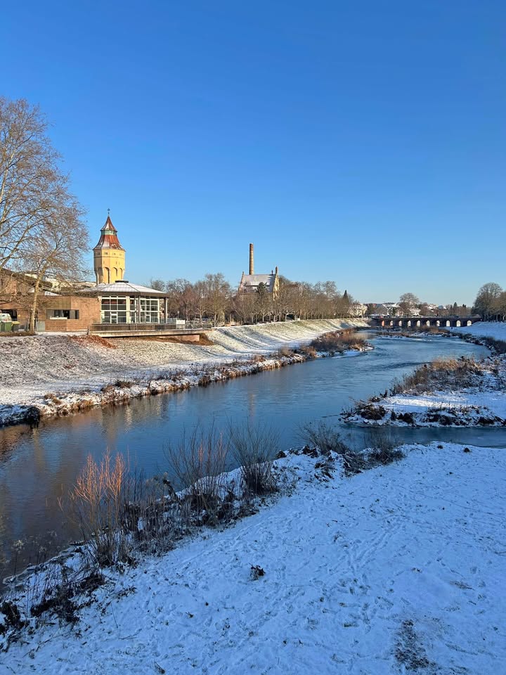 Dieser Alternativtext wurde KI-generiert: Ein Fluss fließt an verschneiten Ufern vorbei. Im Hintergrund sind Gebäude und Bäume unter einem klaren, blauen Himmel zu sehen.