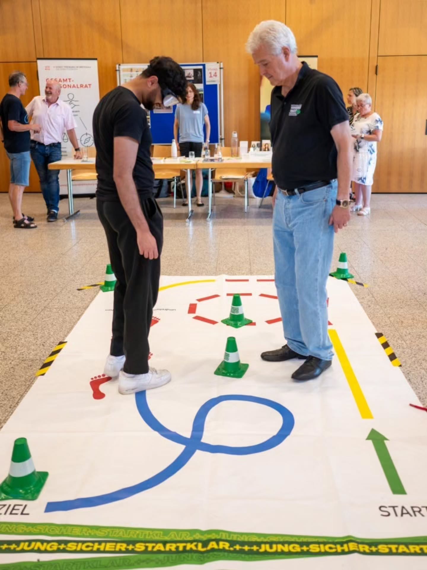 Zwei Personen stehen auf einer großen Bodenmatte mit einem Parcours aus blauen Linien, grünen Kegeln und roten Fußabdrücken; eine trägt eine VR-Brille. Im Hintergrund sind weitere Personen und Informationsstände zu sehen, darunter ein Schild mit „GESAMTPERSONALRAT“. Am Rand der Matte stehen die Wörter „START“ und „ZIEL“. (KI-Alternativtext)