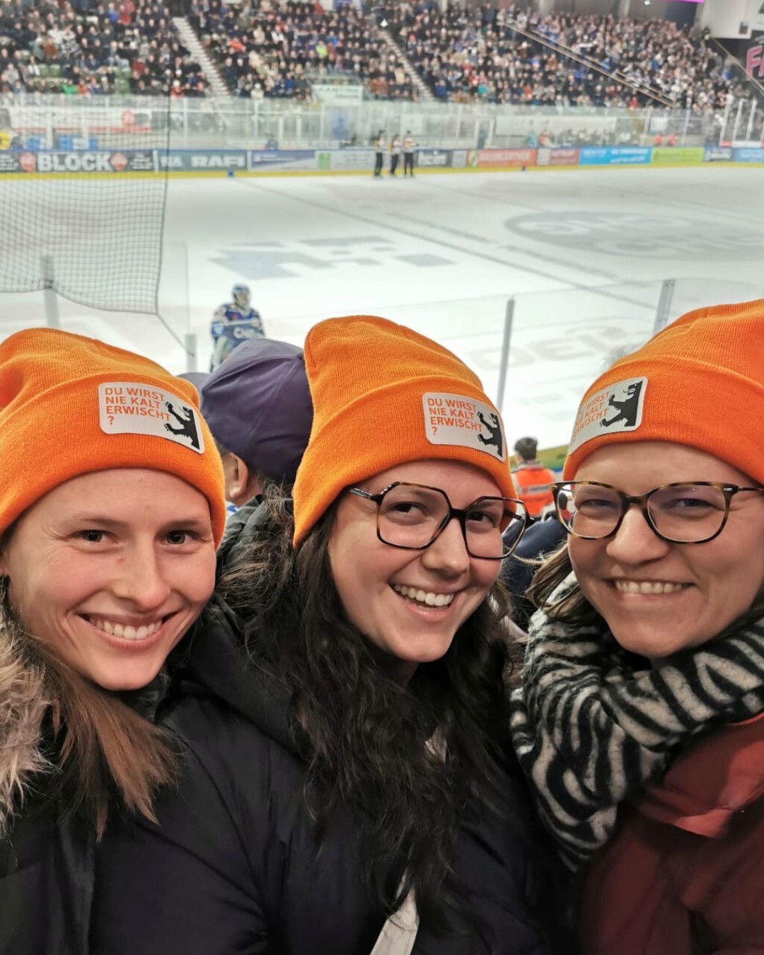 Dieser Alternativtext wurde KI-generiert: Eine Gruppe von Personen mit orangefarbenen Strickmützen lächelt in die Kamera. Im Hintergrund ist ein Eisstadion mit Zuschauern zu sehen.