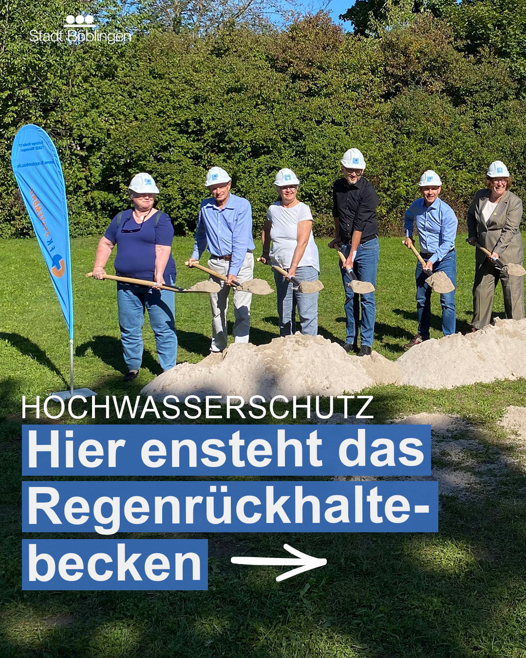 Dieser Alternativtext wurde KI-generiert: Eine Gruppe von Personen mit Helm und Schaufeln steht auf einer Grasfläche vor einer blauen Werbetafel. Auf der Tafel steht in großen Buchstaben 'HOCHWASSERSCHUTZ' und ein Hinweis auf ein Regenrückhaltebecken.