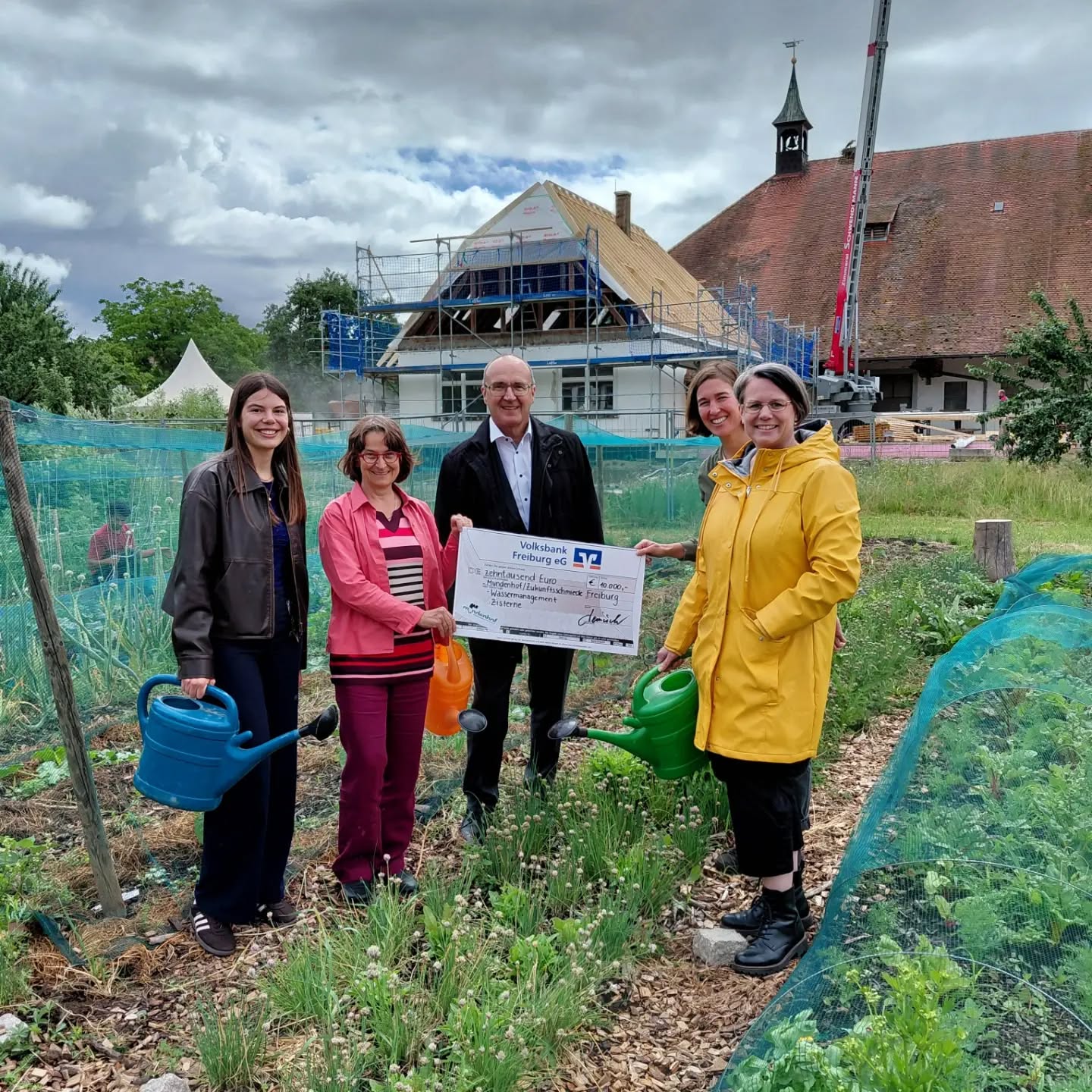 Fünf Personen stehen im Garten vor einem Baugerüst, halten eine große Spendenurkunde der Volksbank Freiburg eG und Gießkannen. Im Hintergrund ist ein Gebäude mit Turm und bewölktem Himmel zu sehen. (KI-Alternativtext)