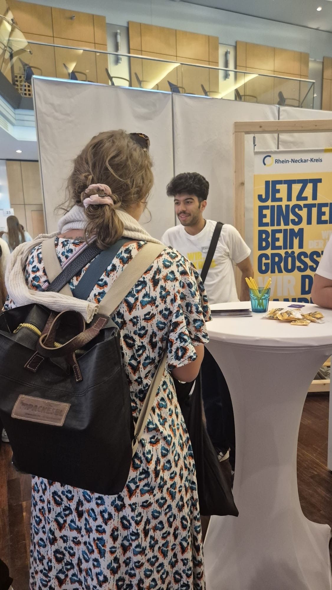 Eine Person mit Rucksack und gemustertem Kleid steht mit dem Rücken zur Kamera und spricht mit einer Person hinter einem weißen Hochtisch. Auf dem Tisch liegen Flyer und ein Becher mit Stiften. Rechts steht ein Banner mit der Aufschrift „Rhein-Neckar-Kreis“ und dem Aufruf „JETZT EINSTEIGEN BEIM GRÖSSTEN“. (KI-Alternativtext)