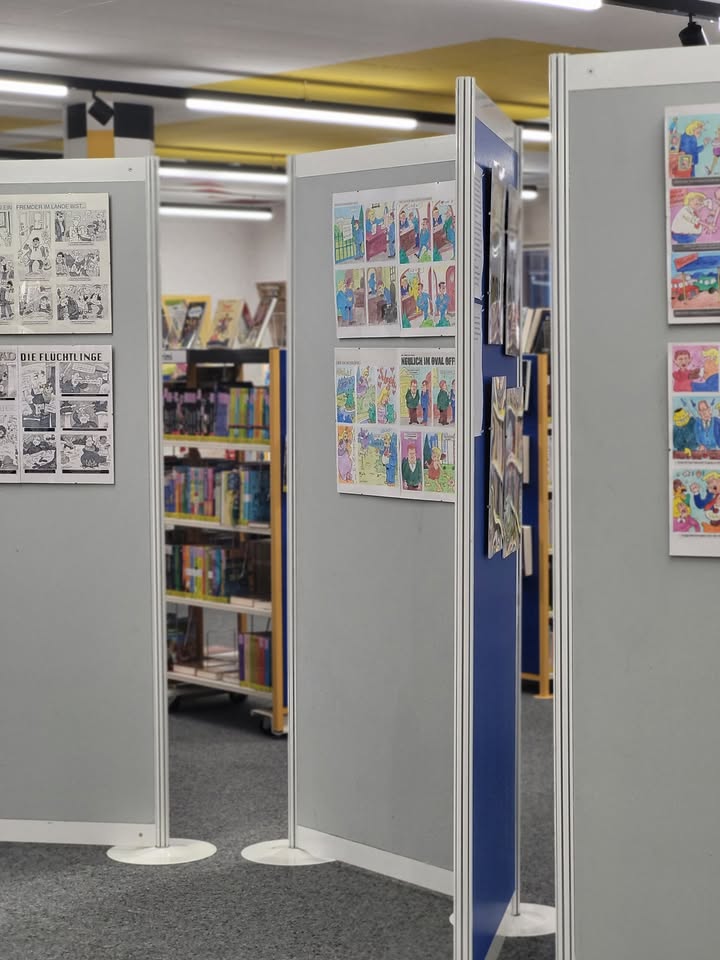 Dieser Alternativtext wurde KI-generiert: Ein Raum mit grauen Stellwänden, an denen Comicseiten und Bilder aufgehängt sind. Im Hintergrund sind Regale mit Büchern und weiteren Comics zu sehen.