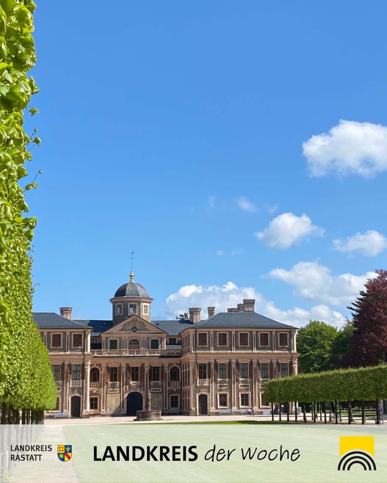 Dieser Alternativtext wurde KI-generiert: Ein großes, helles Gebäude mit vielen Fenstern steht inmitten von Grünanlagen unter einem blauen Himmel. Das Bild ist farbig und zeigt eine architektonisch beeindruckende Fassade.