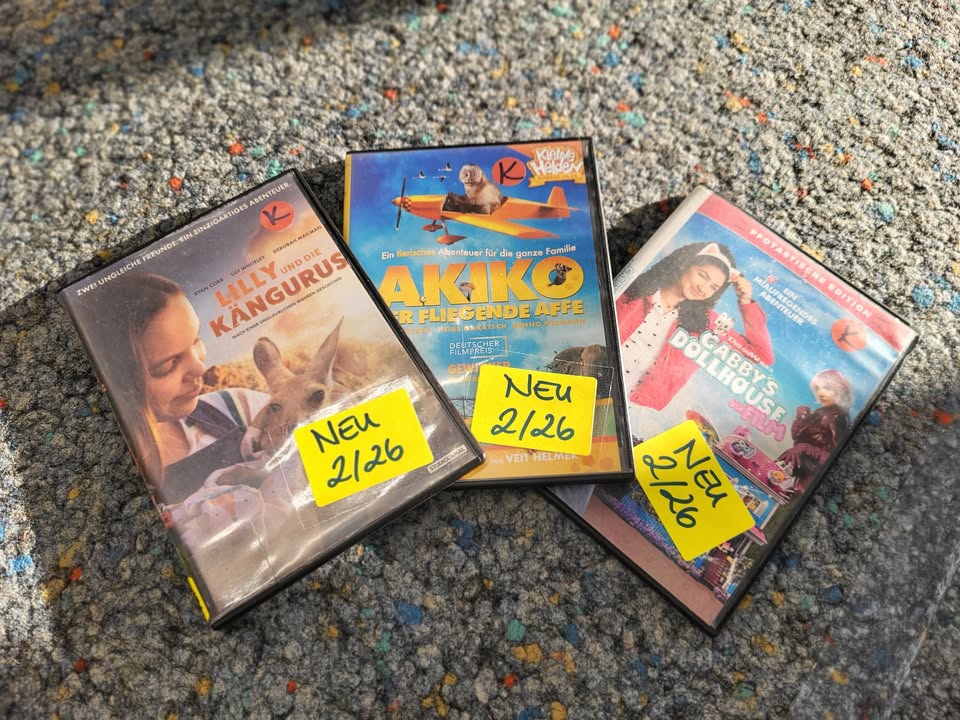 Dieser Alternativtext wurde KI-generiert: Mehrere DVD-Hüllen liegen übereinander auf einem strukturierten, hellbraunen Untergrund. Einige Hüllen haben gelbe Aufkleber mit Zahlen darauf.