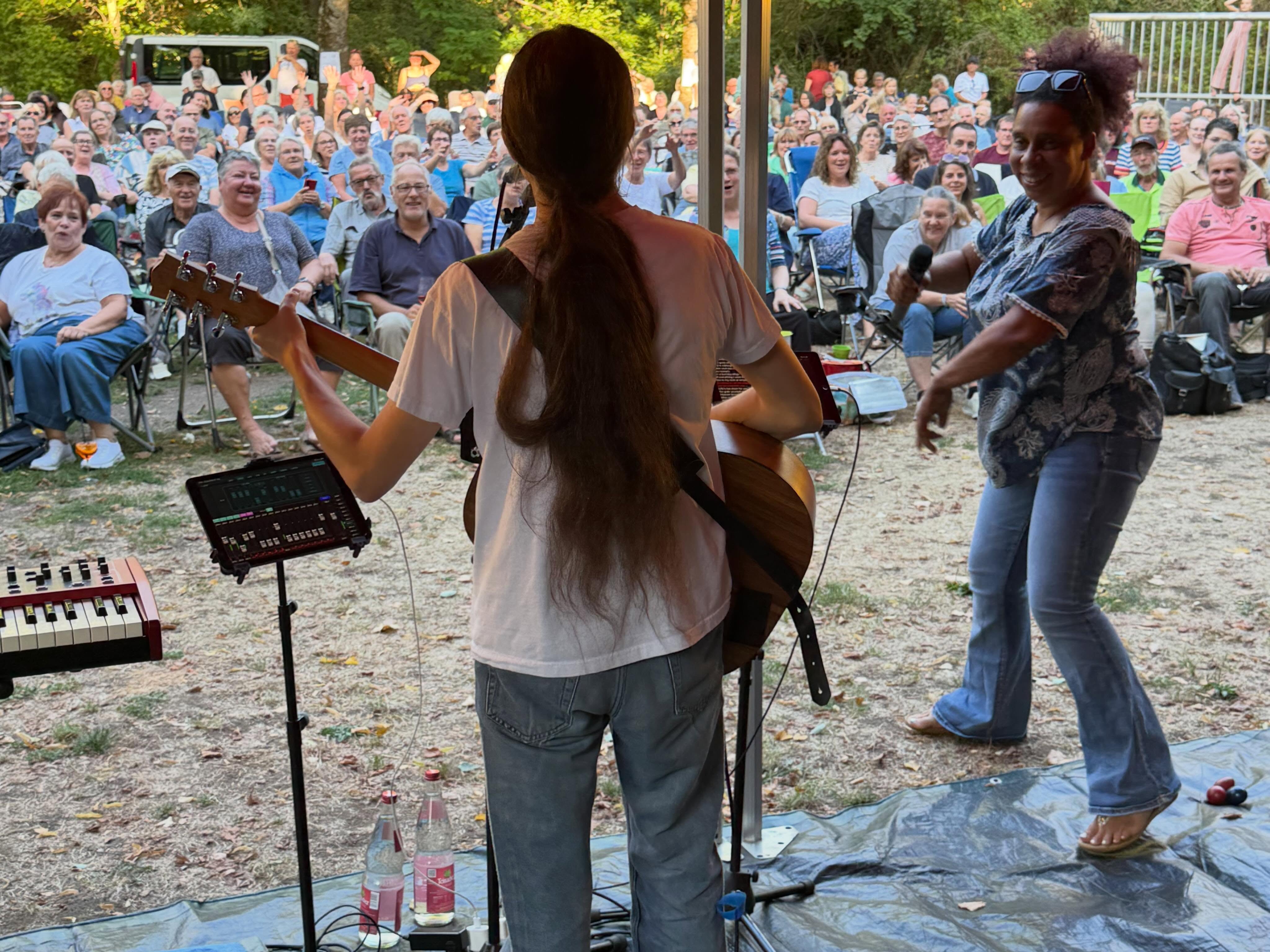 Zwei Personen treten auf einer kleinen Bühne im Freien auf, während ein großes Publikum auf Stühlen auf einer Wiese zuschaut. Eine Person mit langem Haar spielt von hinten gesehen eine Akustikgitarre, während eine weitere Person neben ihr in ein Mikrofon singt. Links im Vordergrund stehen ein Keyboard und ein Tablet auf einem Stativ. (KI-Alternativtext)
