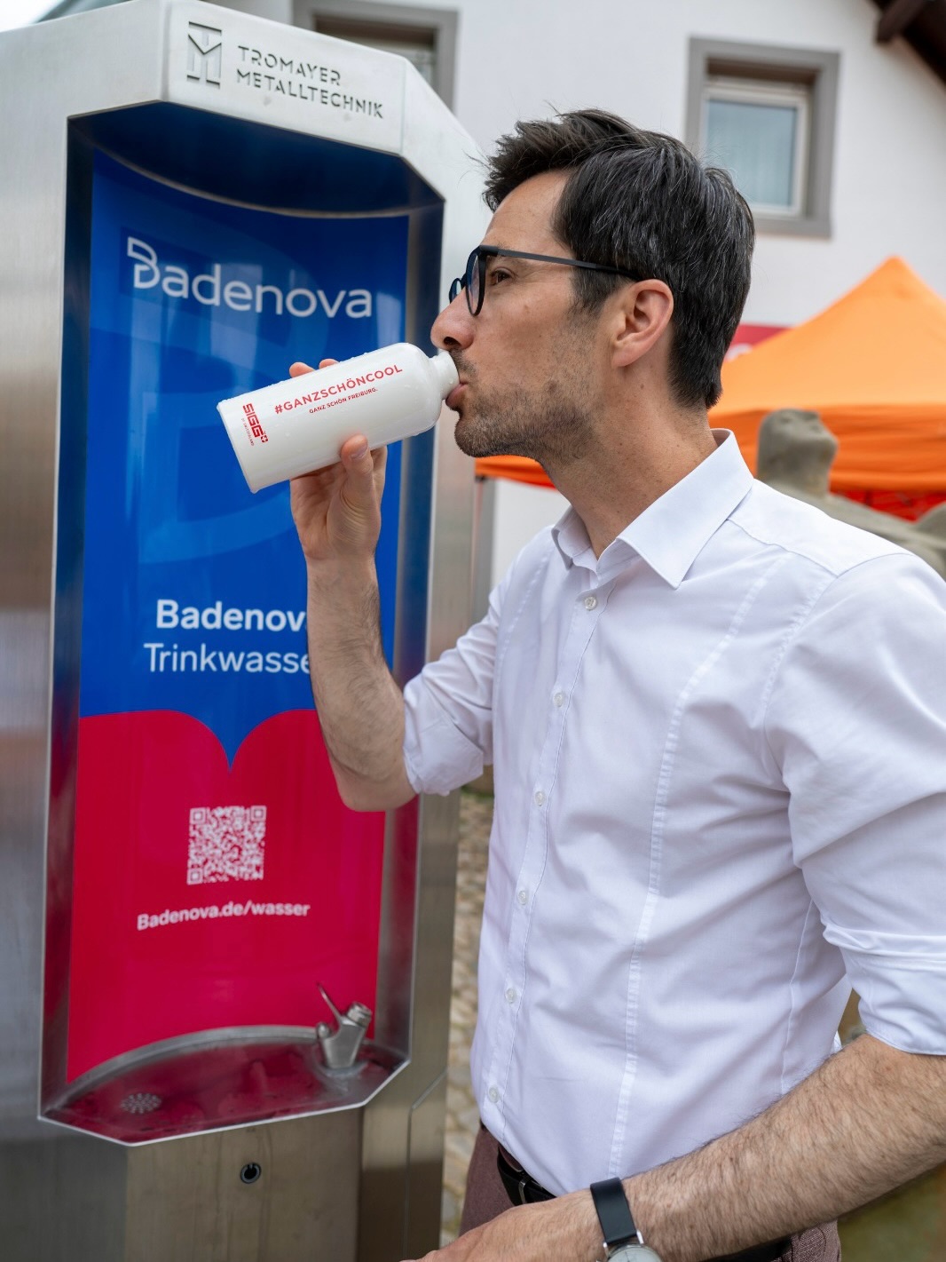Dieser Alternativtext wurde KI-generiert: Eine Person in einem weißen Hemd trinkt aus einer weißen Flasche vor einem metallischen Gerät mit Werbeaufdruck. Im Hintergrund befindet sich eine Wasserquelle.