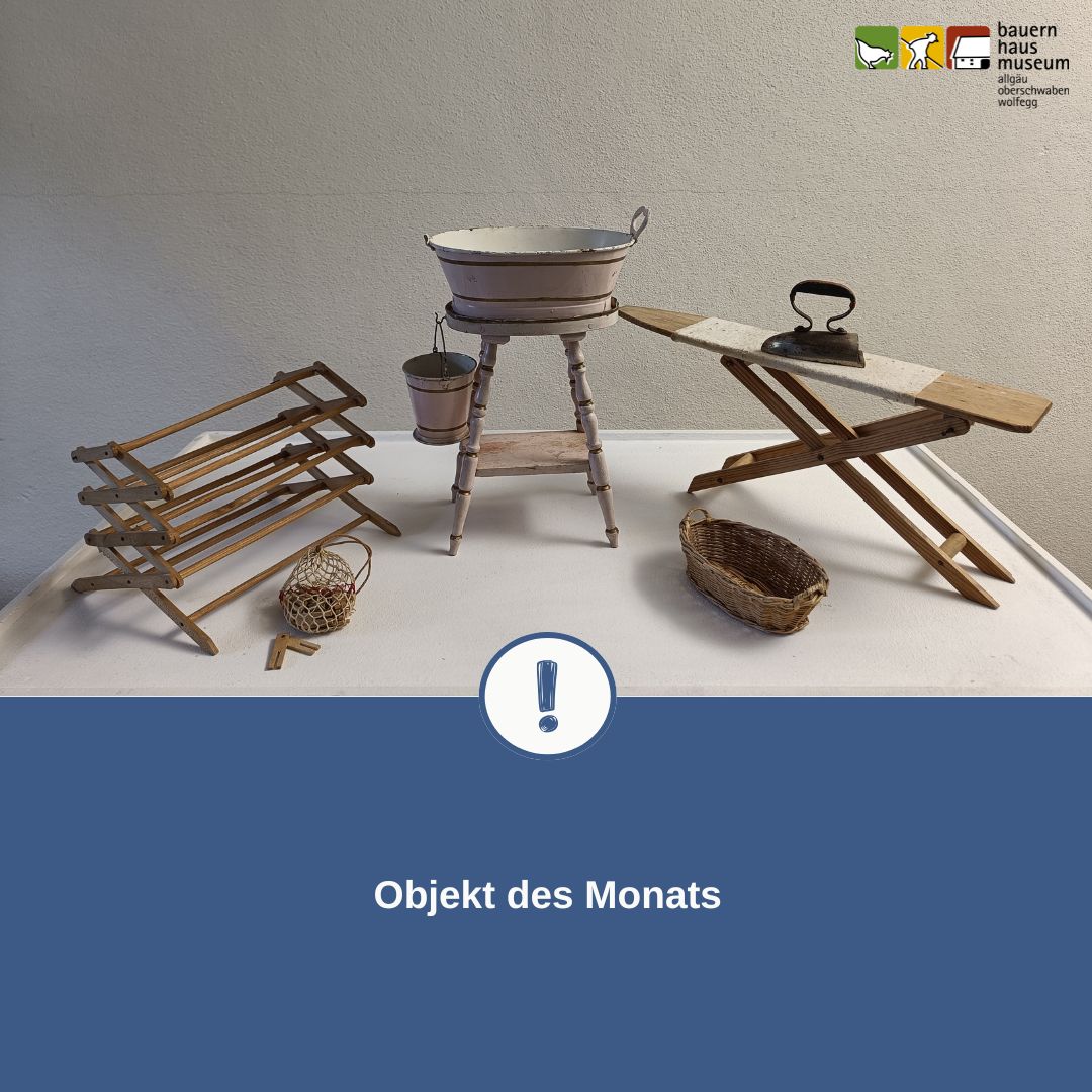 Dieser Alternativtext wurde KI-generiert: Ein Schwarz-Weiß-Foto zeigt verschiedene alte Gegenstände an einer hellen Wand. Dazu gehören ein Wäschebottich, eine Wäschewippe und eine Schubkarre.