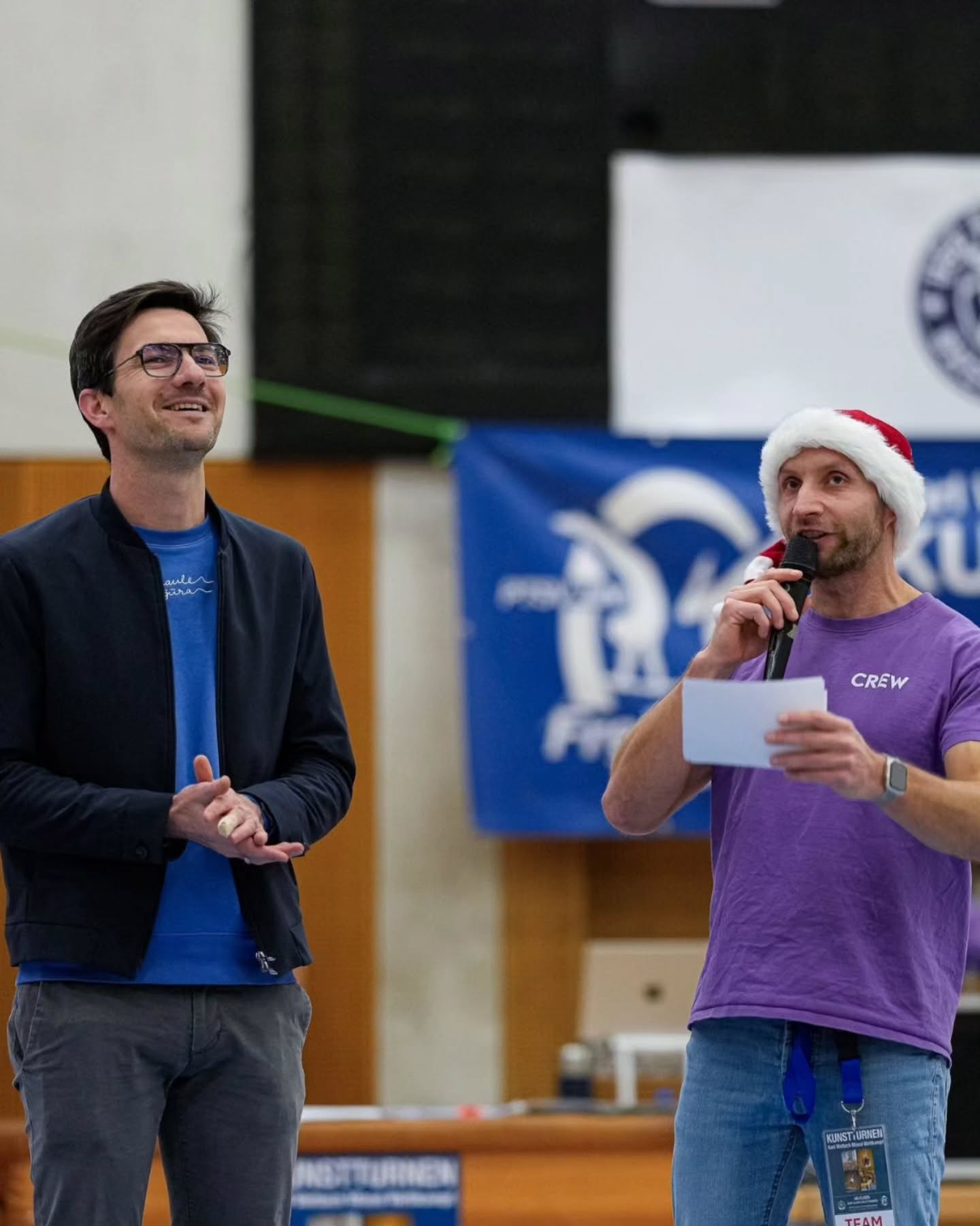 Dieser Alternativtext wurde KI-generiert: Zwei Personen stehen nebeneinander vor einem Banner. Eine Person trägt eine blaue Jacke, die andere einen Weihnachtsmannhut und hält ein Mikrofon.