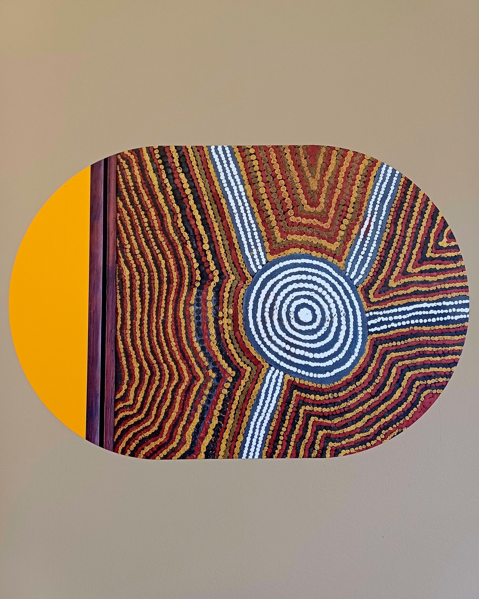 Ein Gemälde im Stil der australischen Aboriginal-Kunst hängt an einer hellbeigen Wand. Links dominiert ein großer gelber Kreis, gefolgt von einem dunklen vertikalen Streifen. Der rechte Bereich zeigt ein komplexes Muster aus Punkten in Rot, Gelb, Weiß und Blau, das sich um ein zentrales, blau-weißes Konzentrisches Kreismuster rankt. (KI-Alternativtext)