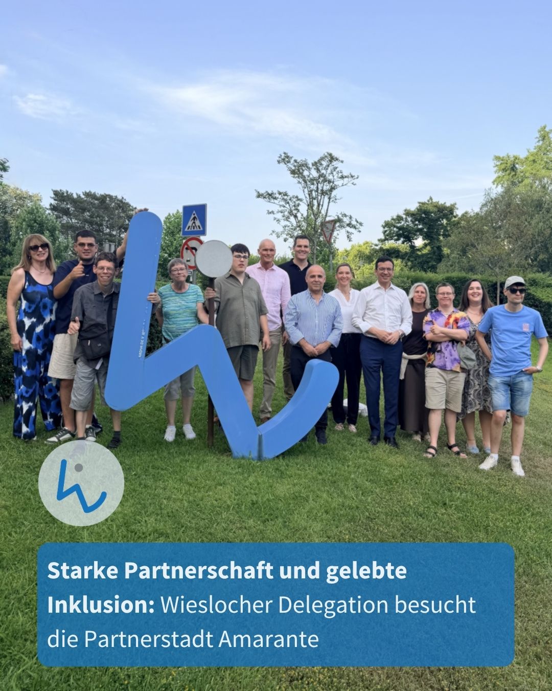 Eine Gruppe von Personen steht im Freien auf einer Wiese und hält gemeinsam ein großes, blaues Schriftzeichen „W“. Ein blauer Textkasten am unteren Rand enthält die Aufschrift „Starke Partnerschaft und gelebte Inklusion: Wieslocher Delegation besucht die Partnerstadt Amarante“ sowie ein Logo mit einem stilisierten W. (KI-Alternativtext)