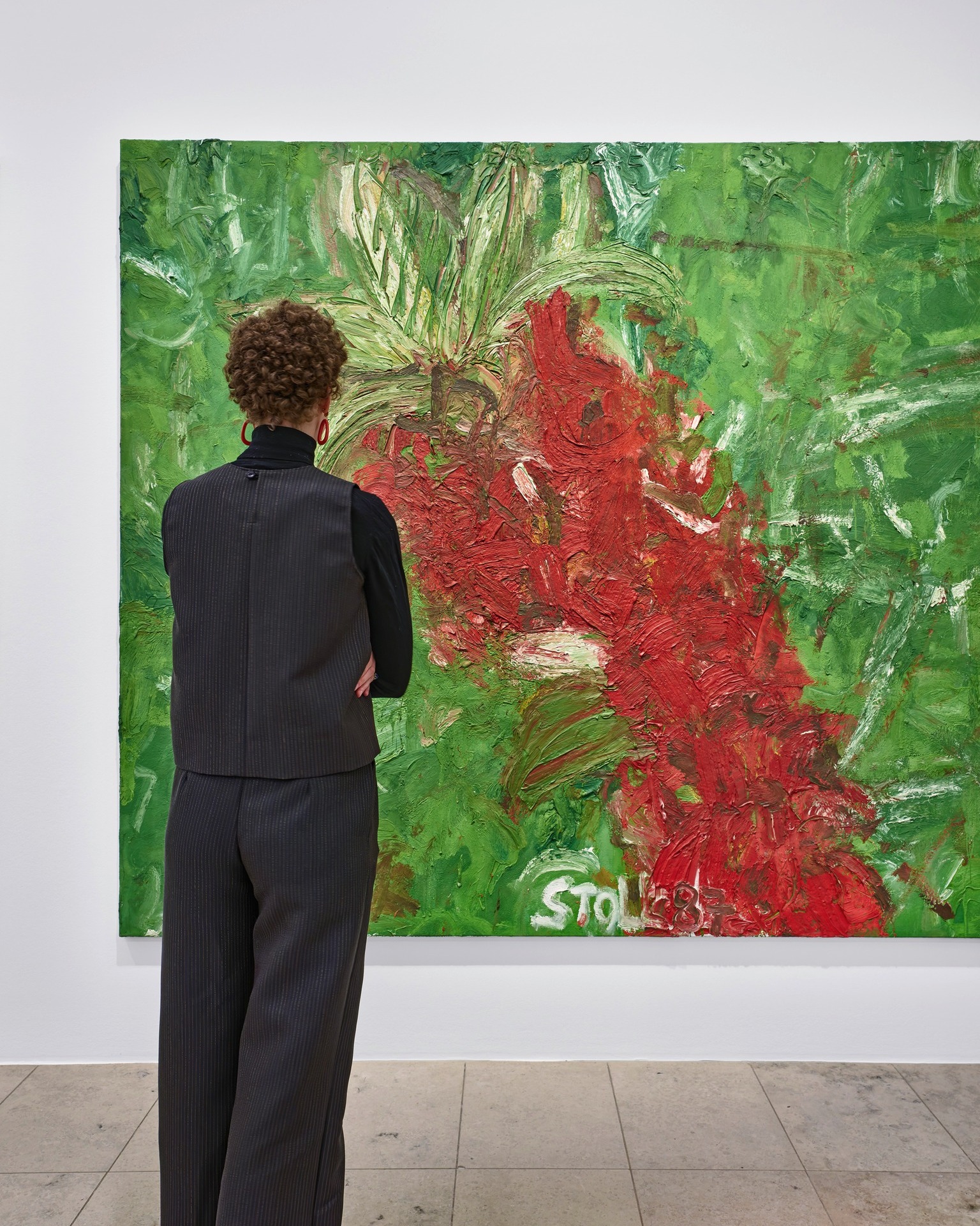 Dieser Alternativtext wurde KI-generiert: Eine Person steht mit dem Rücken zum Betrachter vor einem großen, farbenfrohen Gemälde. Das Bild zeigt rote und grüne Farbtupfer auf einer hellen Wand in einem Raum.