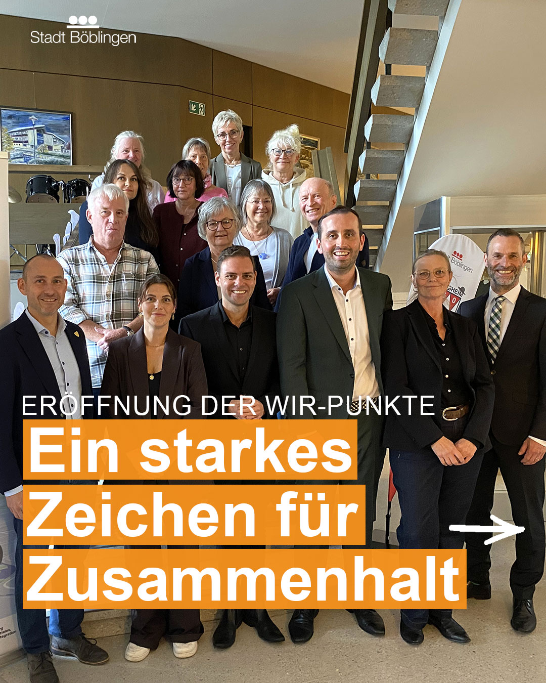 Dieser Alternativtext wurde KI-generiert: Eine Gruppe von Menschen steht in einem Innenraum vor einer Treppe und lächelt in die Kamera. Im Vordergrund befindet sich ein Banner mit Text und Symbolen.