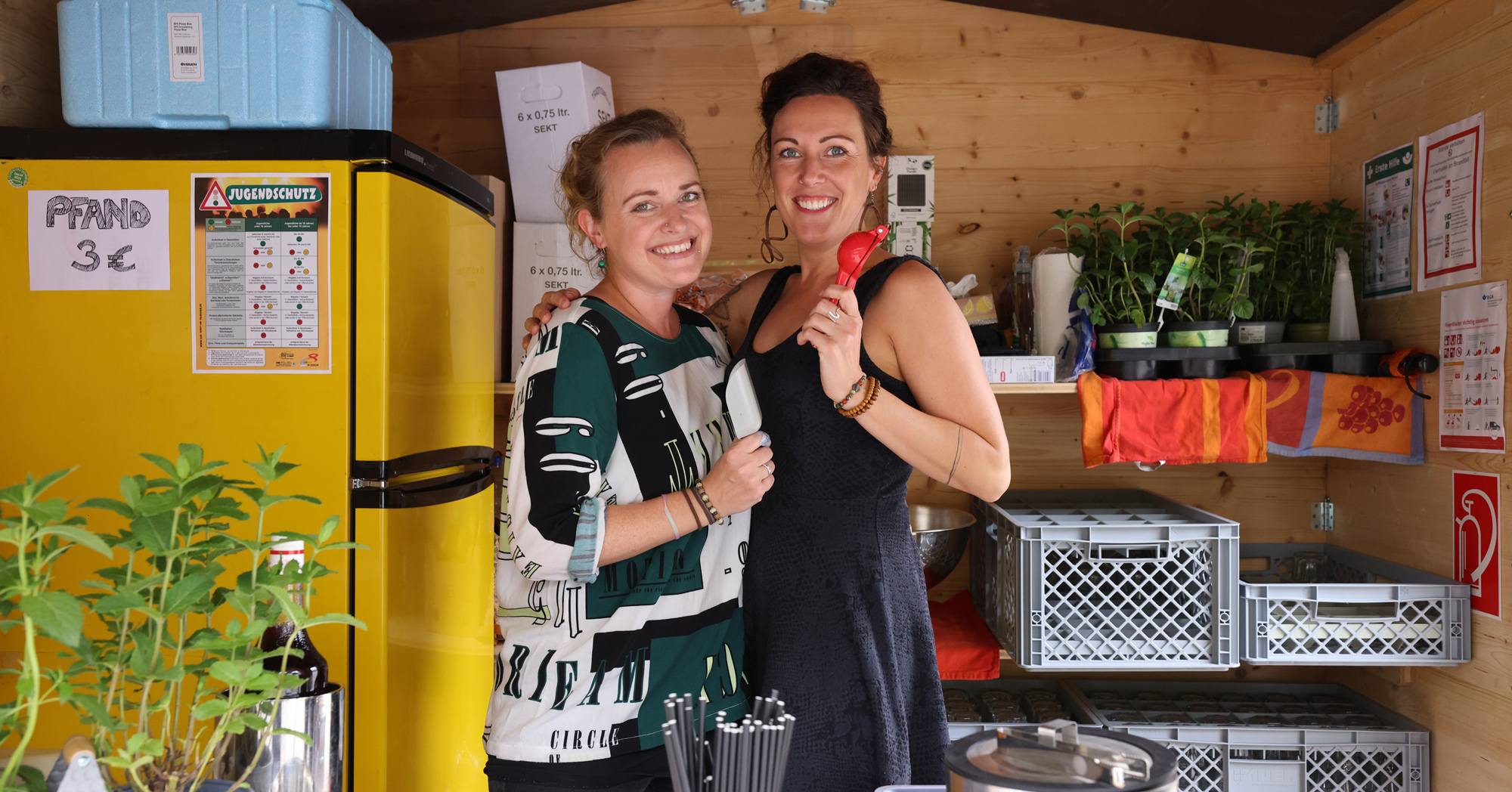 Zwei Frauen stehen in einem kleinen Laden. Im Hintergrund ist ein Kühlschrank und Regale zu sehen.
