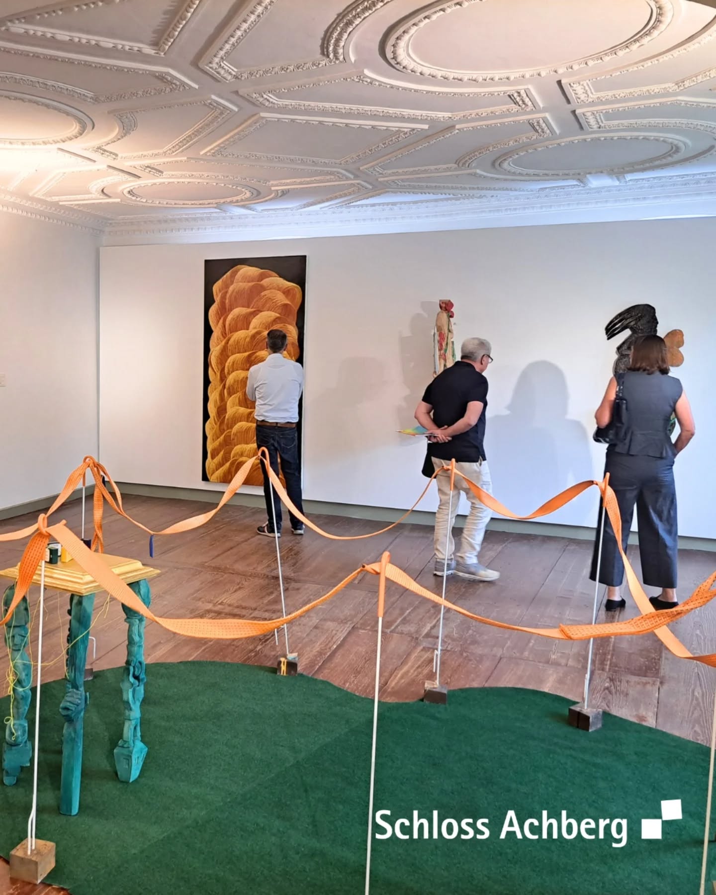Innenaufnahme eines Kunstausstellungsraumes mit Personen, die Kunstwerke an den Wänden betrachten. Der Raum hat eine dekorative Decke und eine grüne Bodenfläche vor einigen Kunstwerken.