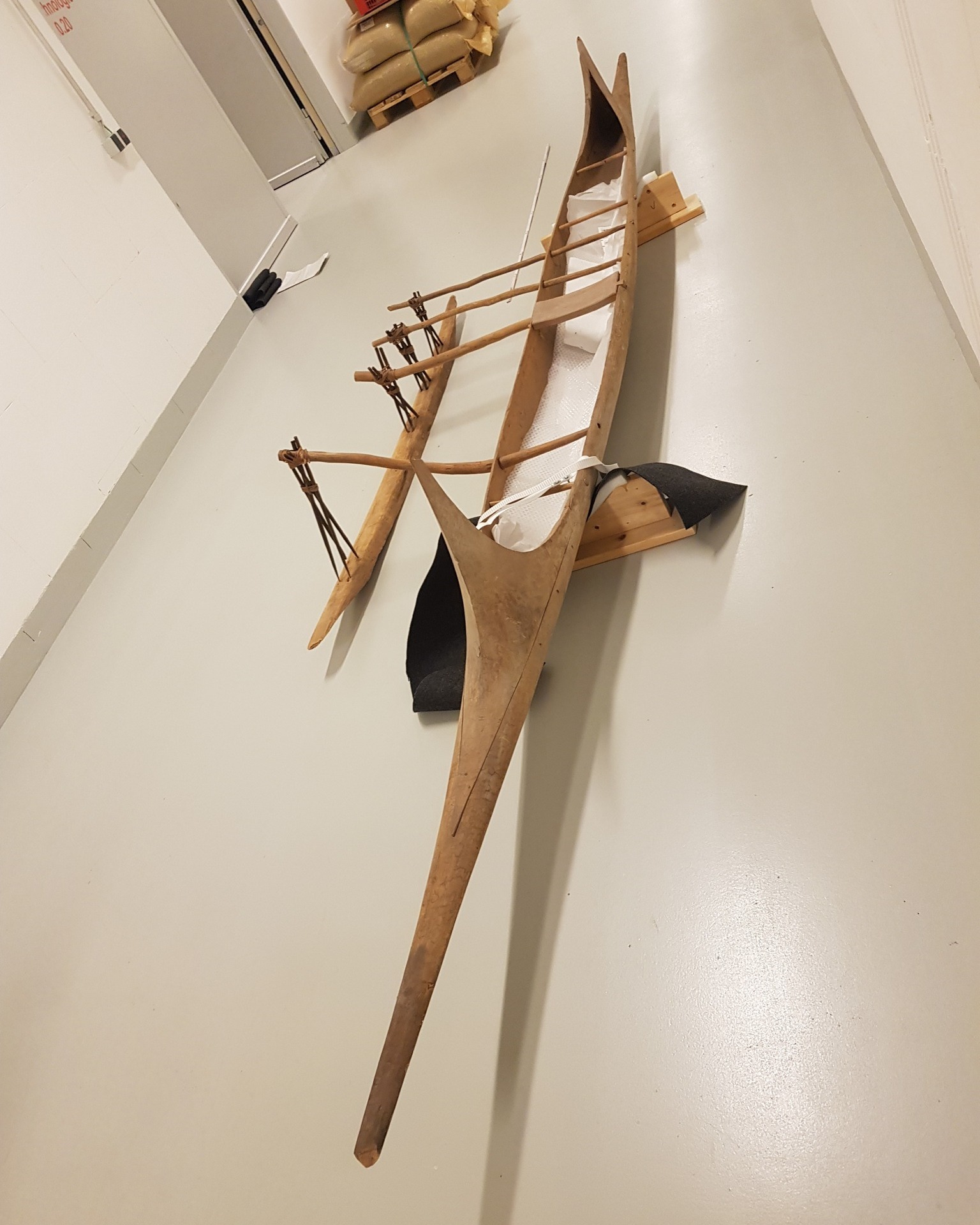 Dieser Alternativtext wurde KI-generiert: Ein hölzernes Kanu, vermutlich ein Modell, lehnt an einer weißen Wand. Das Objekt zeigt eine traditionelle Bauweise mit einem schmalen Rumpf und einem gerippten Rahmen.