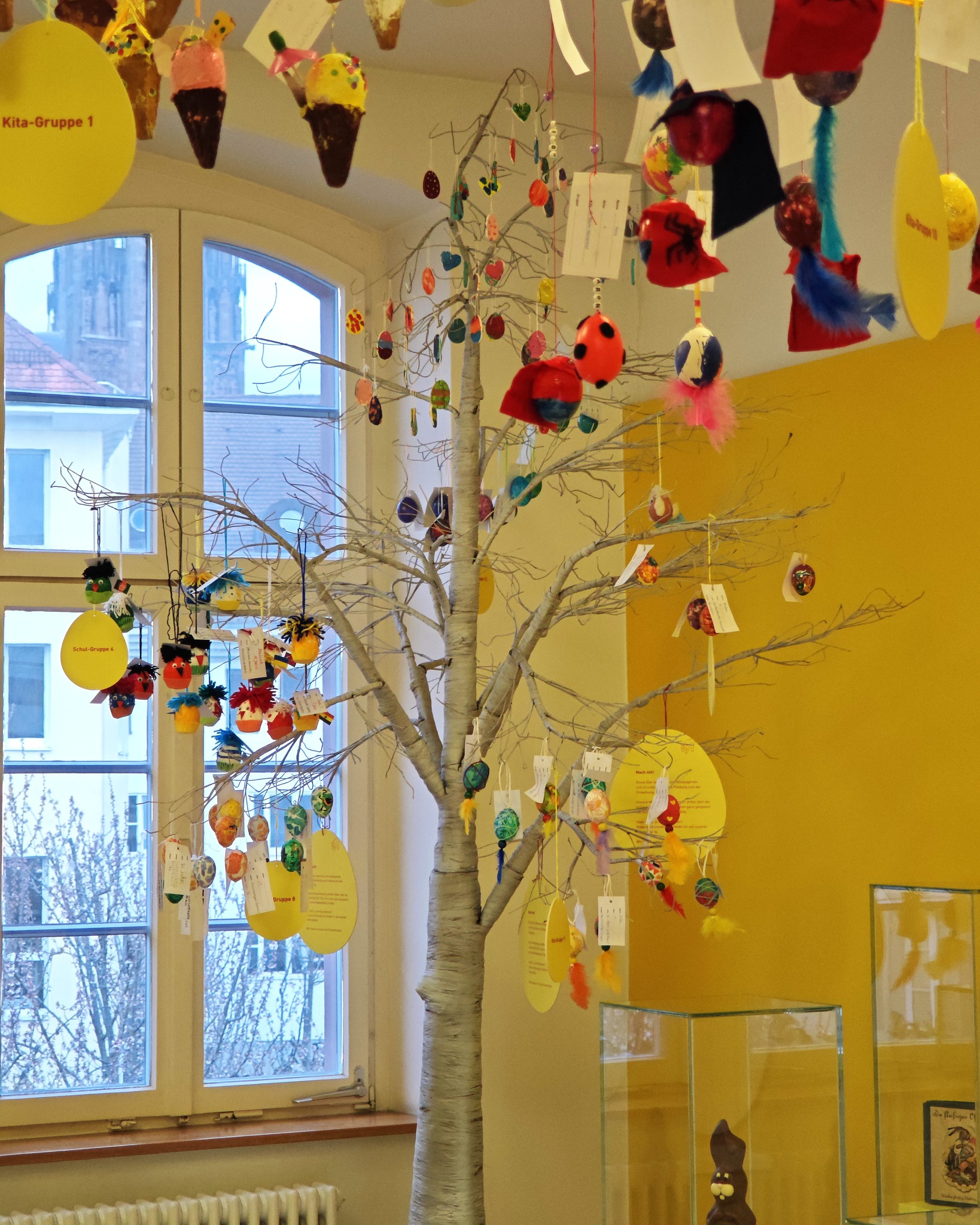 Dieser Alternativtext wurde KI-generiert: Ein Innenraum mit einem Baum, an dem viele bunte Dekorationen hängen. Im Hintergrund ist ein Fenster mit Blick auf Bäume zu sehen.