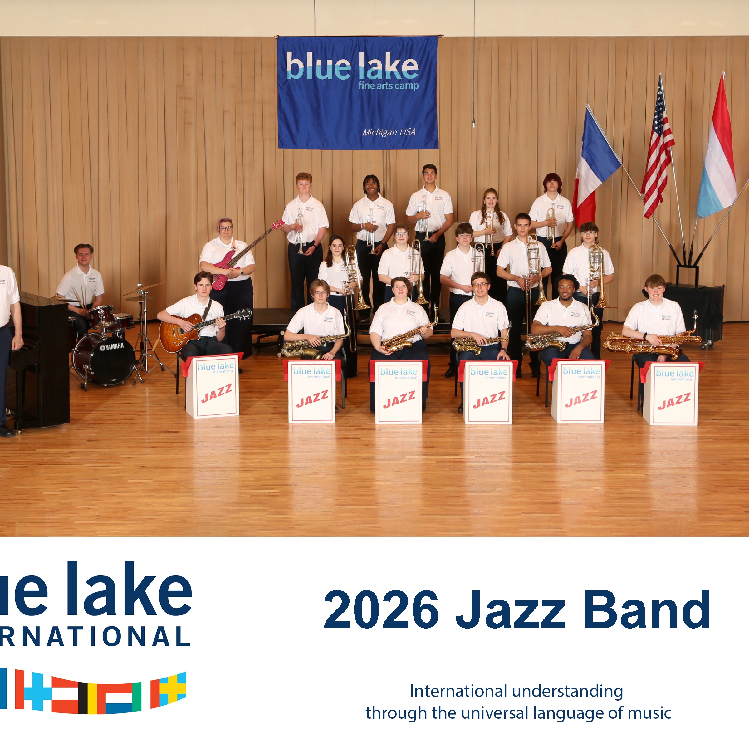 Gruppenfoto einer Jazzband vor Holzwand mit blauem „blue lake fine arts camp“-Banner und drei Flaggen; Musiker in weißen Hemden mit Instrumenten, vorne Schilder mit „JAZZ“. Unten Text: „2026 Jazz Band – International understanding through the universal language of music“. (KI-Alternativtext)