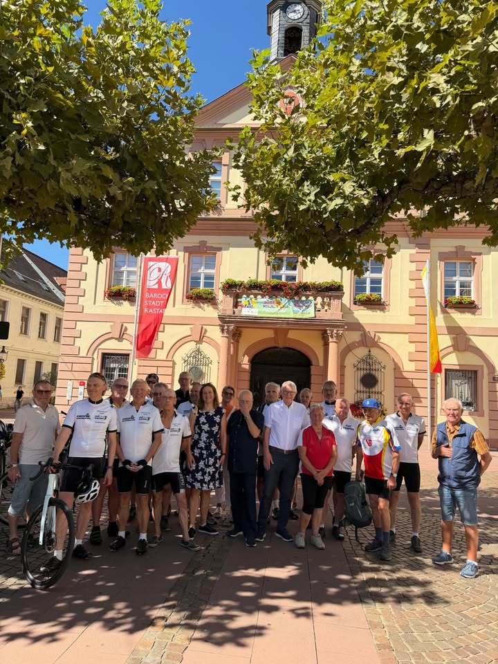 Eine große Gruppe von Personen posiert vor einem hellen Gebäude mit Uhrturm. Viele tragen weiße T-Shirts, eine Person hält ein Fahrrad. Eine rote Fahne trägt die Aufschrift "BAROCK STADT RASTATT". (KI-Alternativtext)