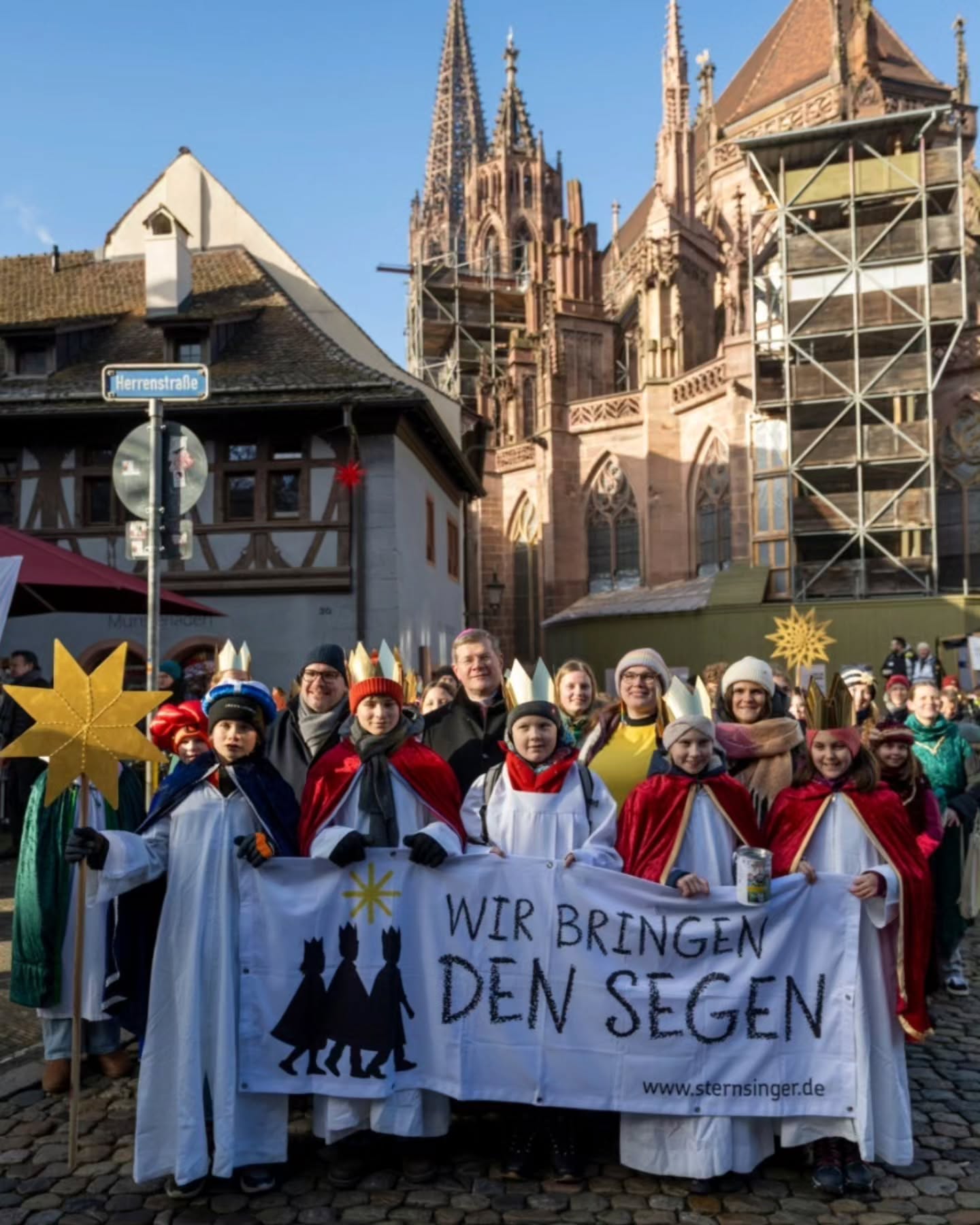 Dieser Alternativtext wurde KI-generiert: Eine Gruppe von Personen steht auf einem Pflasterplatz vor historischen Fachwerkhäusern und einer Kirche. Sie halten Sterne und ein Banner mit einer Botschaft.