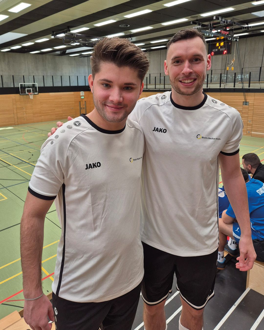 Dieser Alternativtext wurde KI-generiert: Zwei Personen in hellen Sporttrikots mit dunklen Akzenten stehen nebeneinander und schauen in die Kamera. Im Hintergrund ist eine Sporthalle mit einer bunten Wandgestaltung zu sehen.