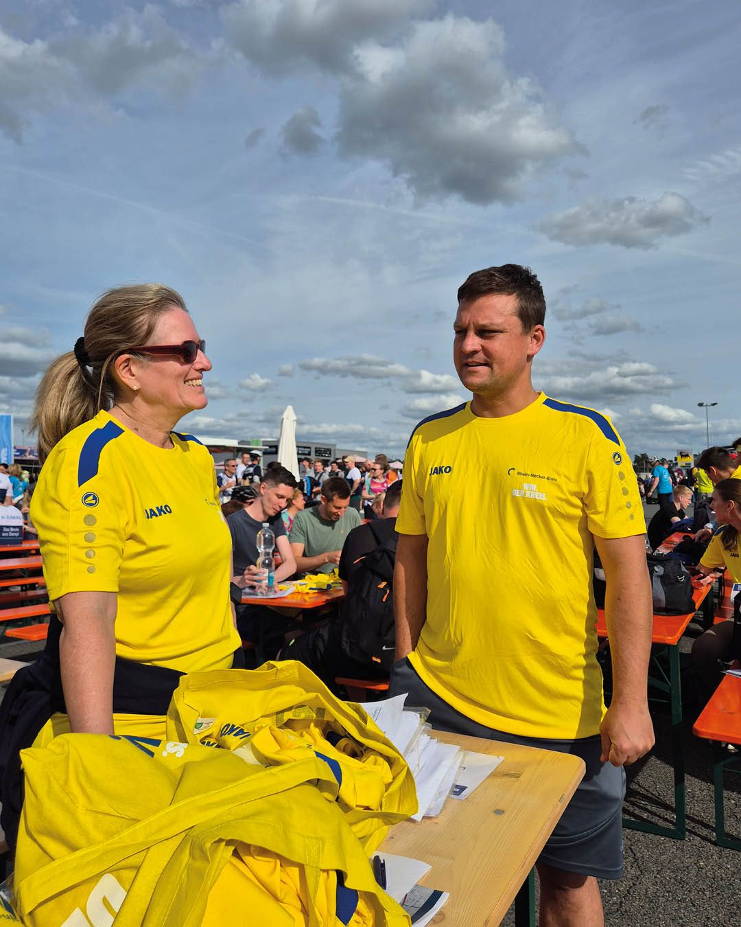 Zwei Personen in gelben JAKO-Shirts stehen im Freien an einem Tisch mit weiteren gelben Shirts und Papieren. Im Hintergrund sitzen Menschen an Tischen unter einem bewölkten Himmel. (KI-Alternativtext)