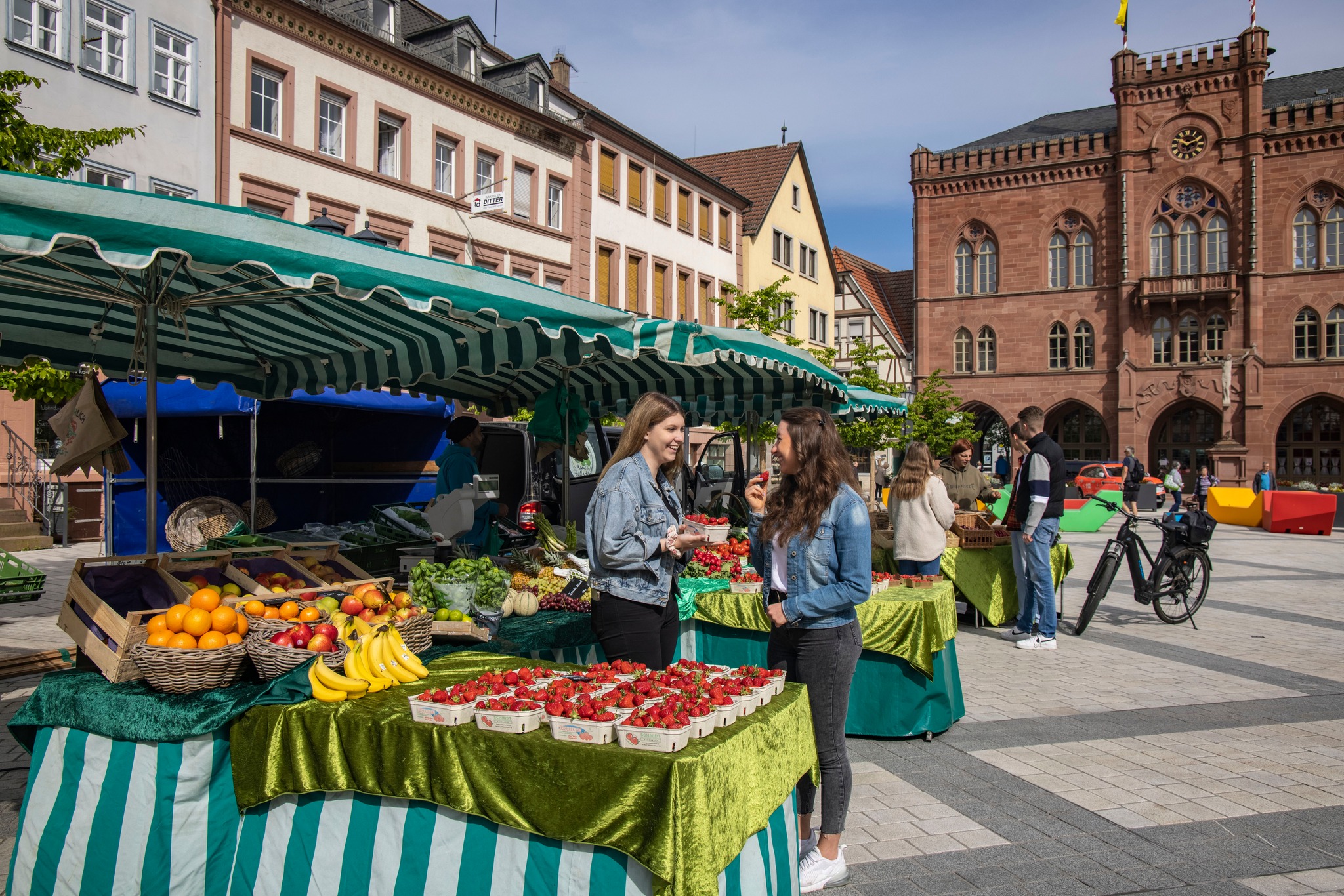 Zwei Personen unterhalten sich an einem Marktstand mit verschiedenen Früchten. Im Hintergrund sieht man weitere Marktstände und Gebäude.