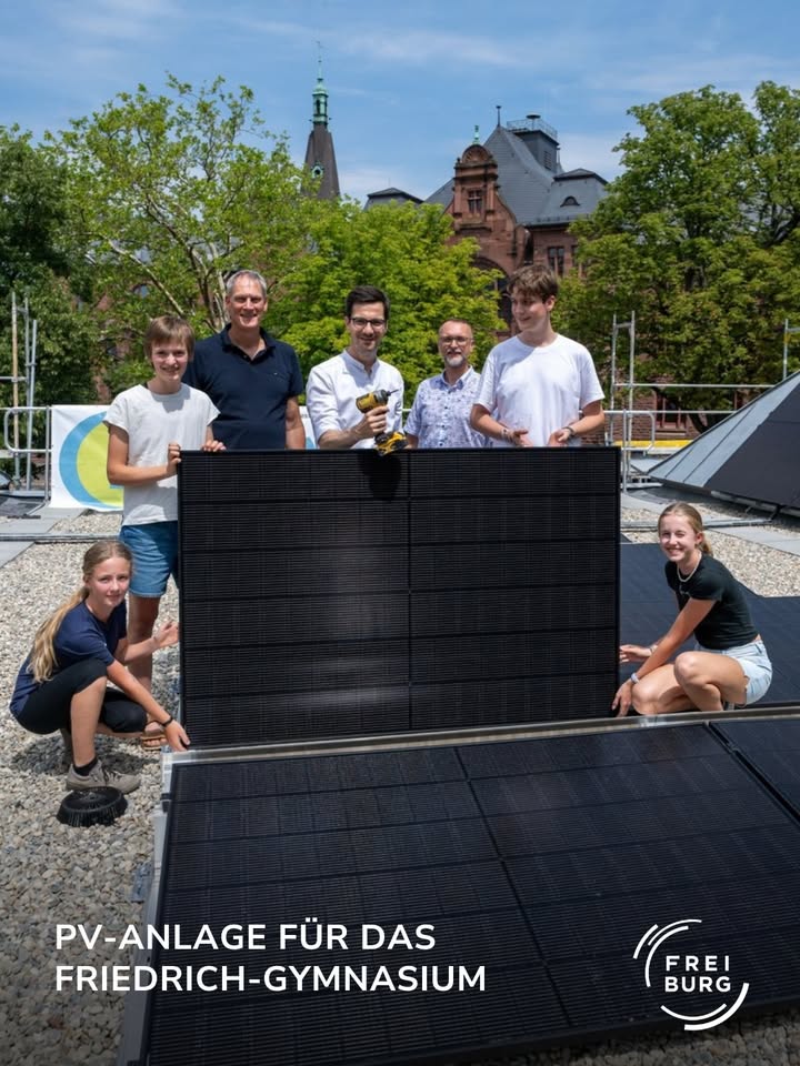 Eine Gruppe von Personen steht und kniet auf einem Dach um ein aufgestelltes Solarmodul herum, während ein weiteres Modul flach im Vordergrund liegt. Eine Person hält eine Bohrmaschine. Der Text im Bild lautet „PV-ANLAGE FÜR DAS FRIEDRICH-GYMNASIUM“ sowie das Logo „FREI BURG“. (KI-Alternativtext)